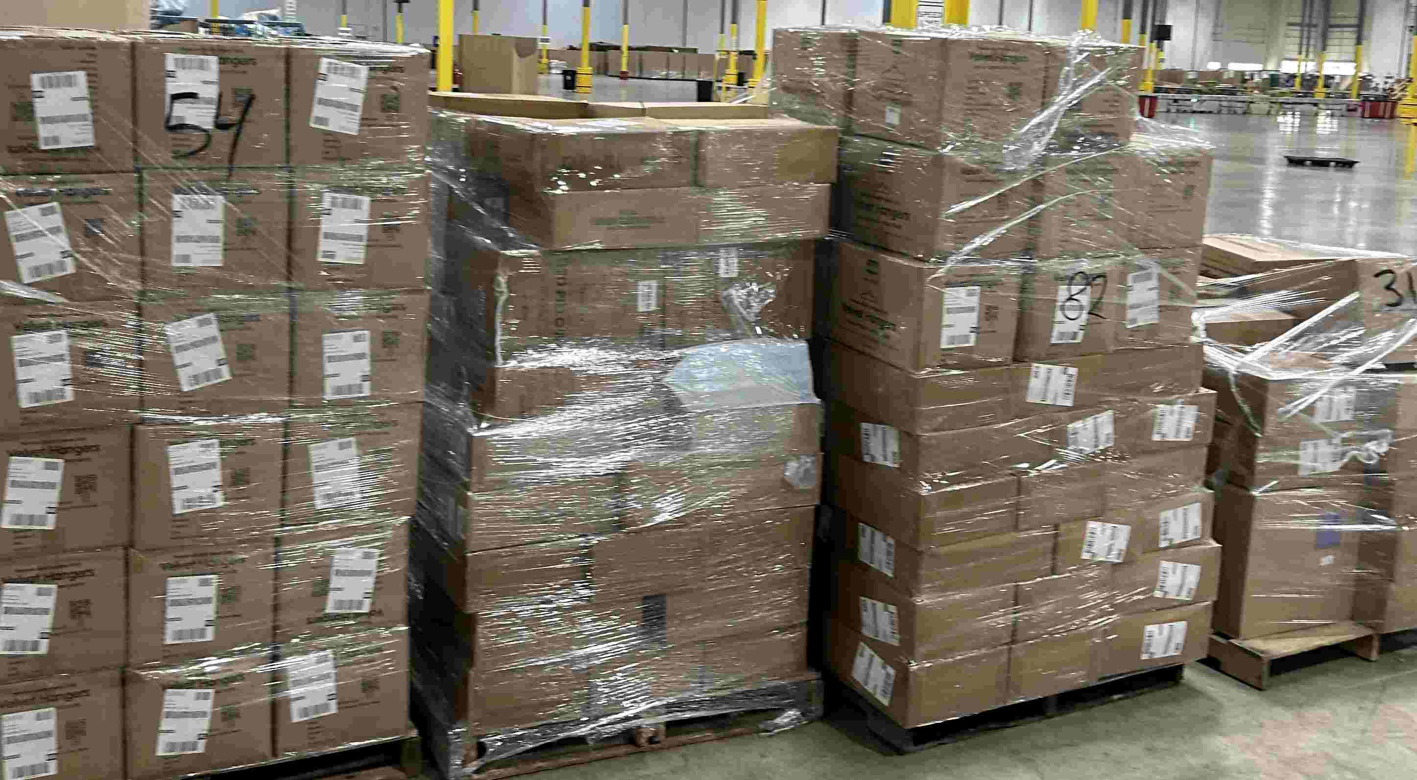 Stacks of cardboard boxes wrapped in plastic on pallets in a warehouse, with shelves and equipment in the background.