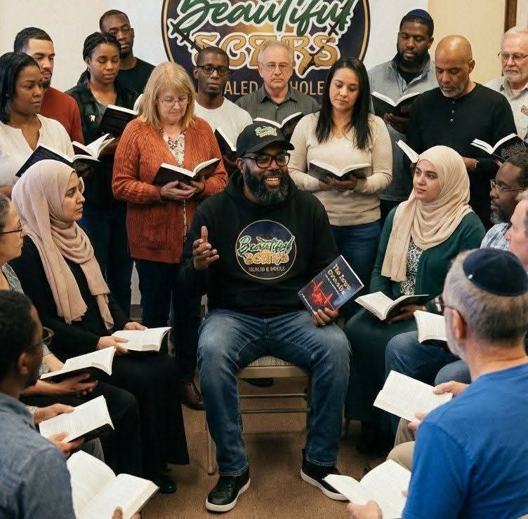 Group of diverse people gathered around a man speaking, holding books, in a room with a sign reading 'Beautiful Voices'.