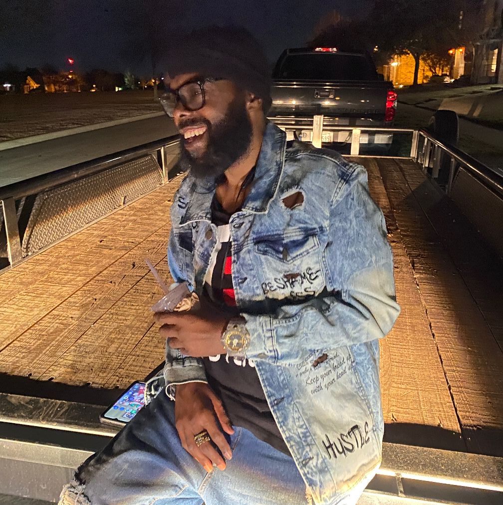 Man with glasses and beard sitting on a wooden boat at night, smiling, wearing a denim jacket and watch.
