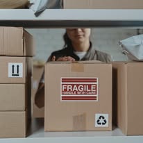 Person behind boxes with a 'Fragile' label, partially visible face, in a warehouse setting.
