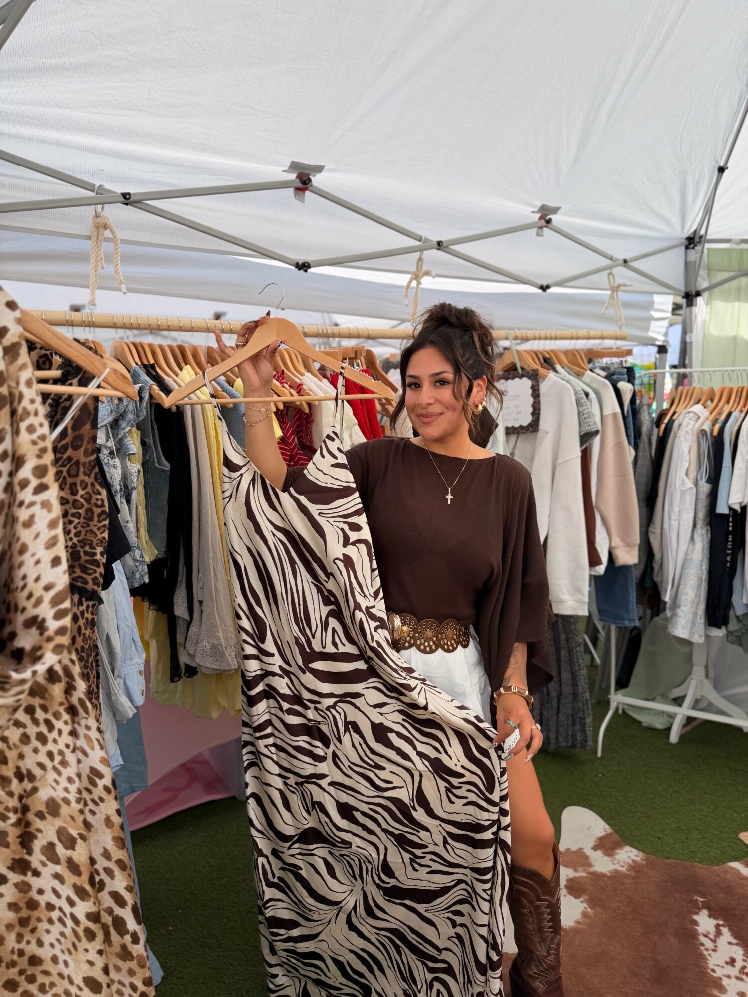 Woman in black top and animal print skirt standing under a tent with clothing racks behind her.