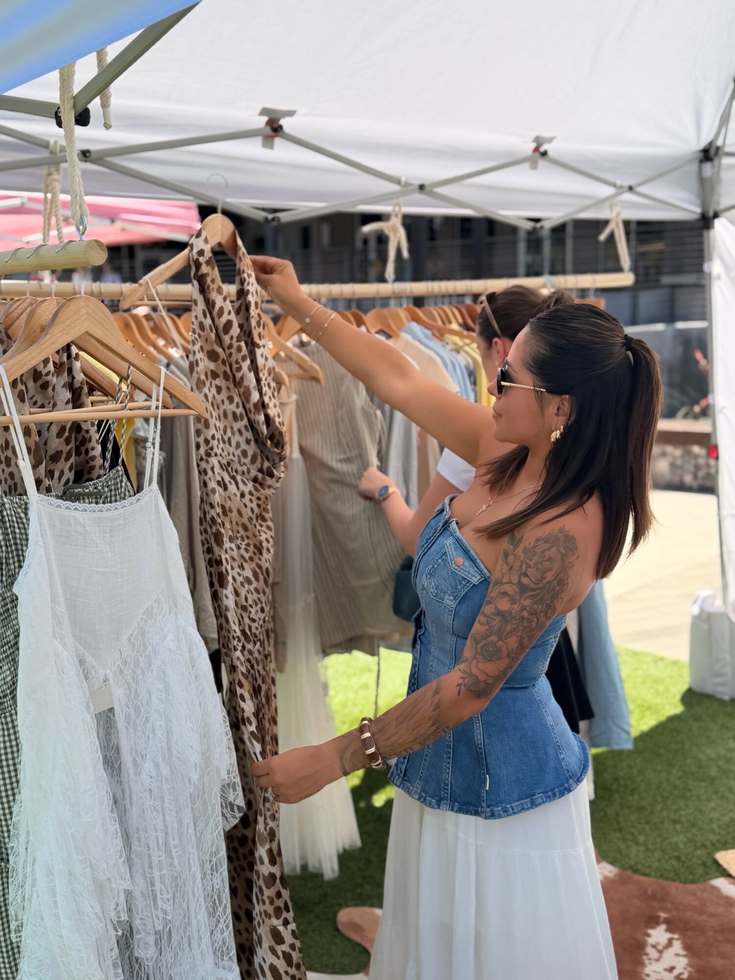 Woman shopping for clothes at outdoor market, browsing animal print and white garments, under a white canopy tent.