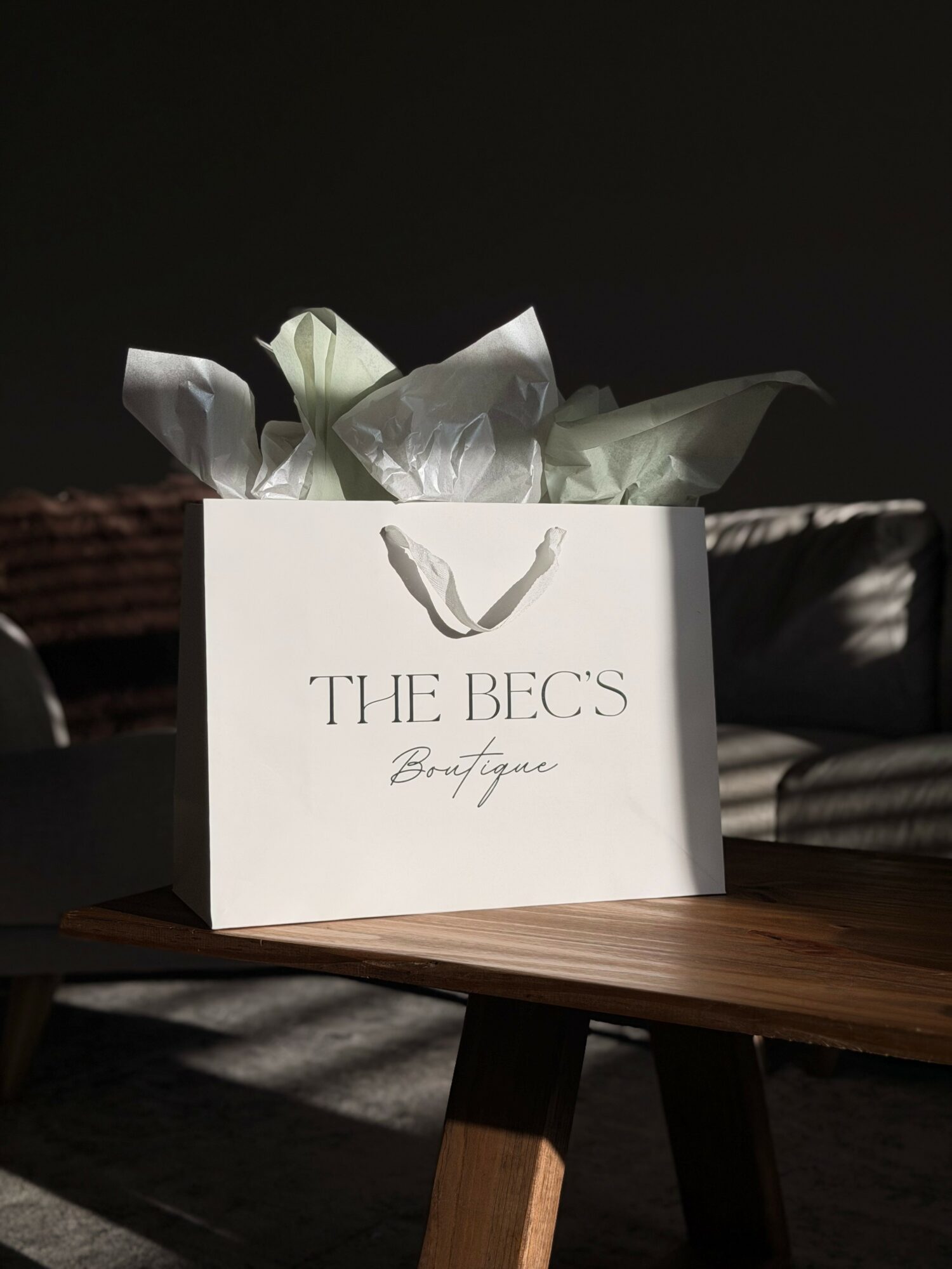 A white shopping bag with tissue paper inside, placed on a wooden table, with a dark background.