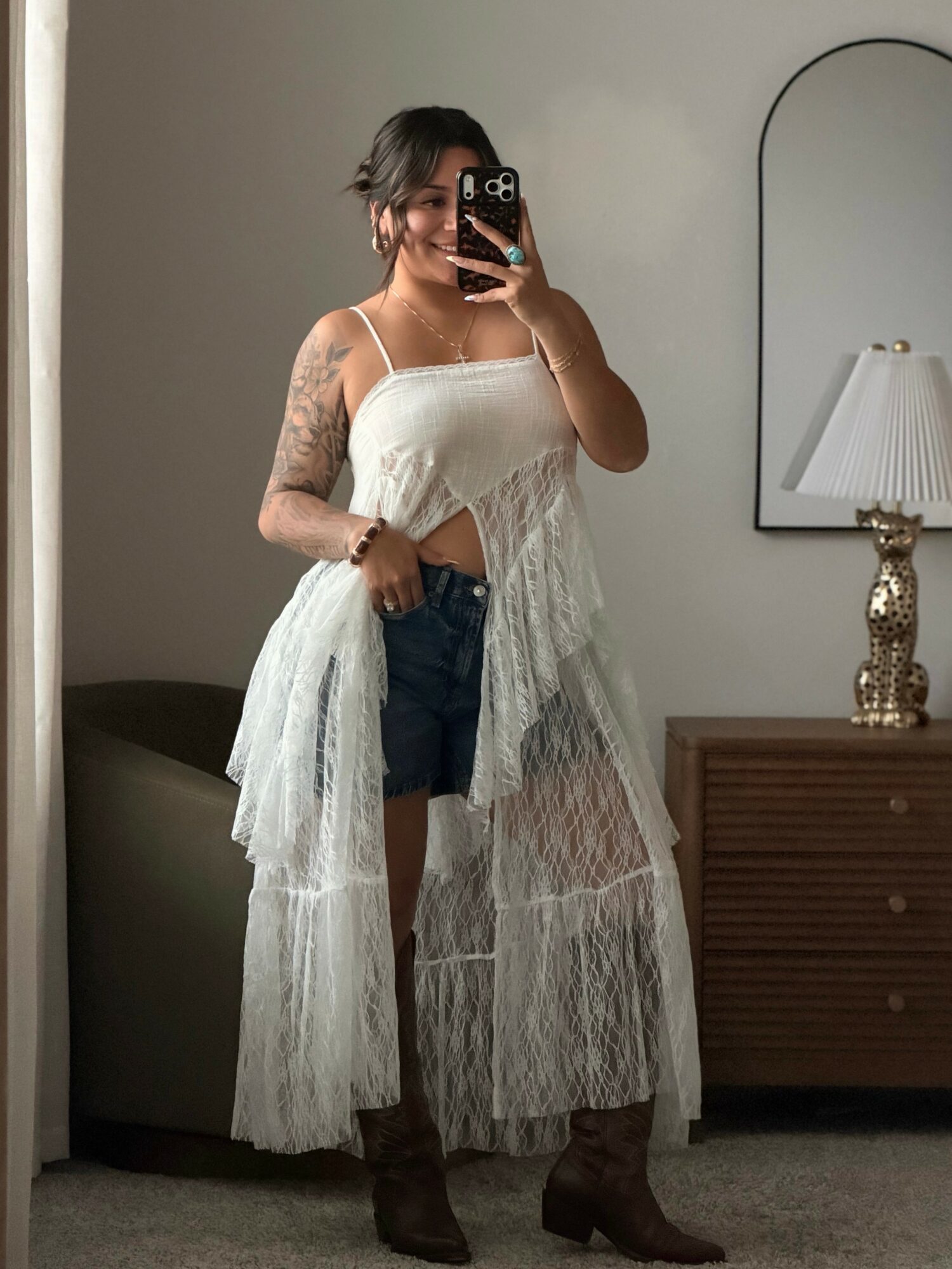 Person taking mirror selfie, wearing a white lace dress over denim shorts, with tattoos on arms, in a room with a dresser and lamp.