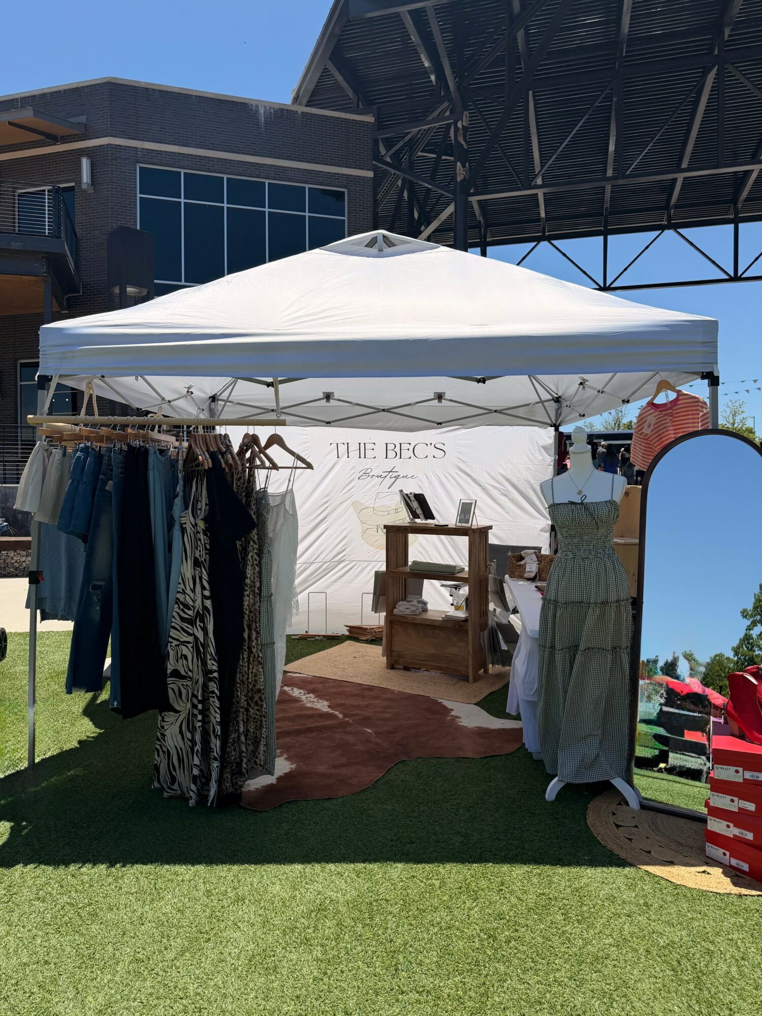 Outdoor market stall with clothing and a mirror under a white canopy, building and blue sky in background.