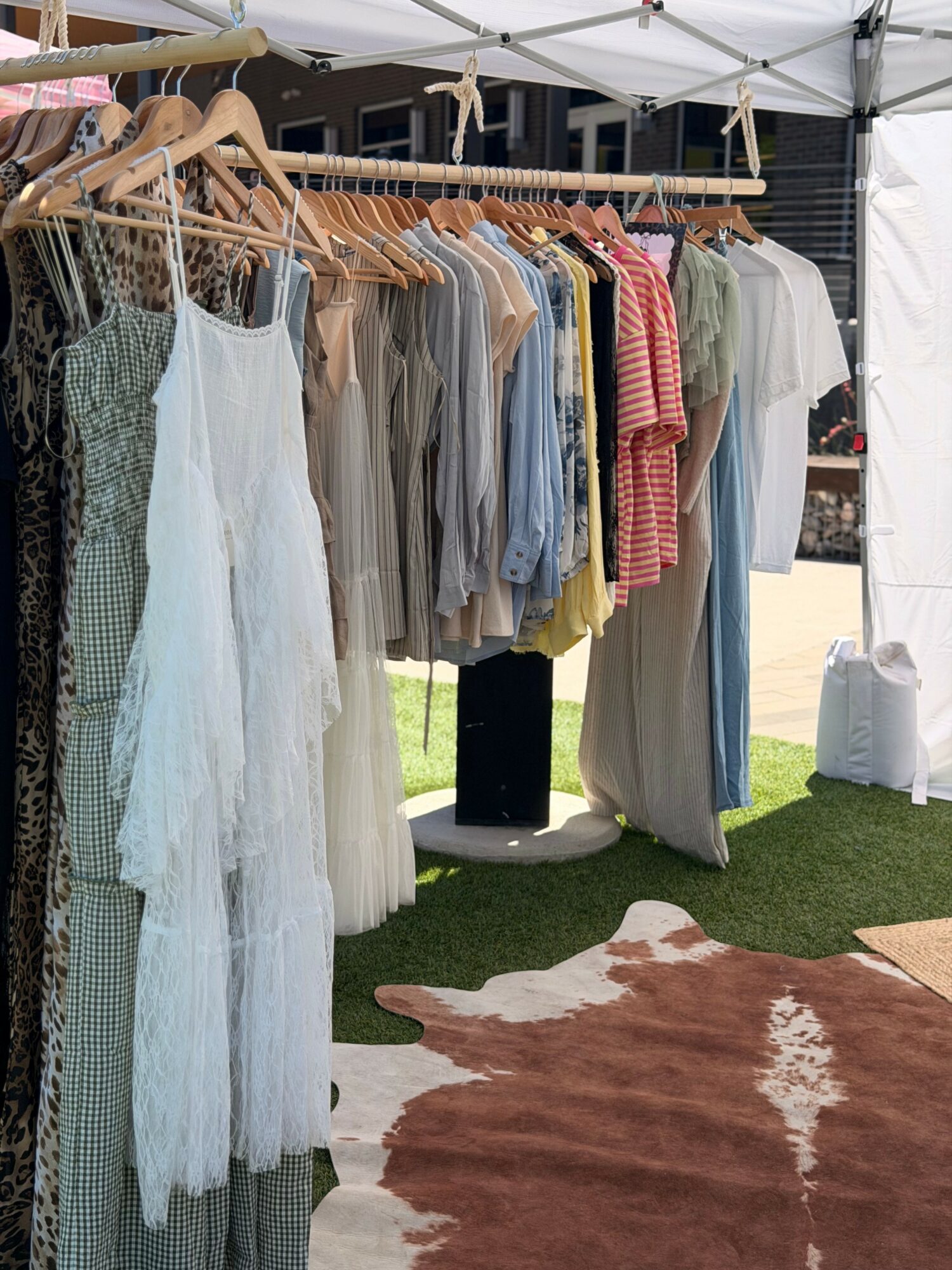 Clothing rack with dresses and shirts under a white canopy, on grass with a cowhide rug nearby.