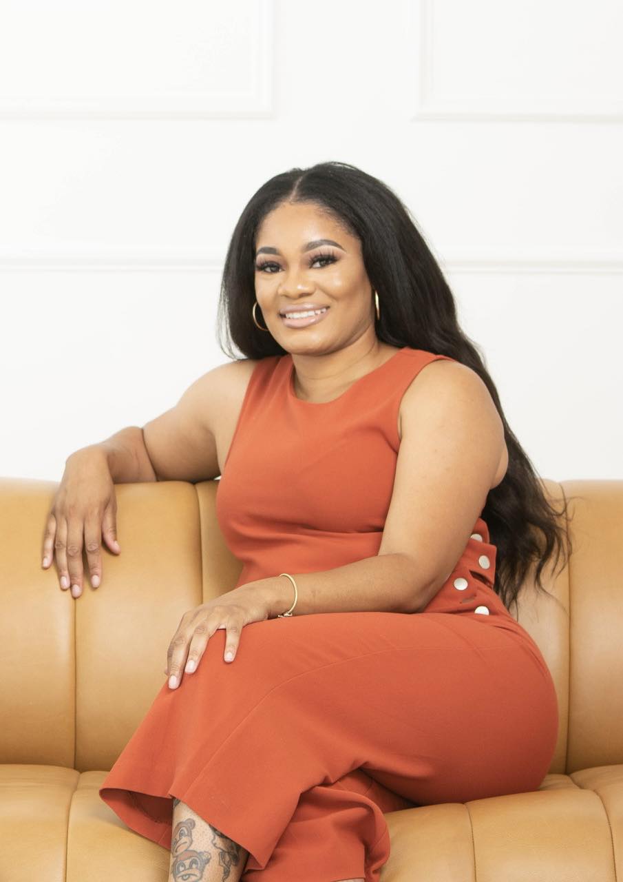 Woman with long dark hair sitting on a tan couch, smiling, wearing an orange sleeveless dress, in a bright room.