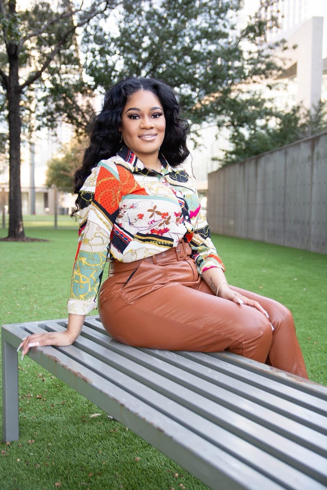 Woman sitting on a park bench outdoors, smiling, with trees and a building in the background.