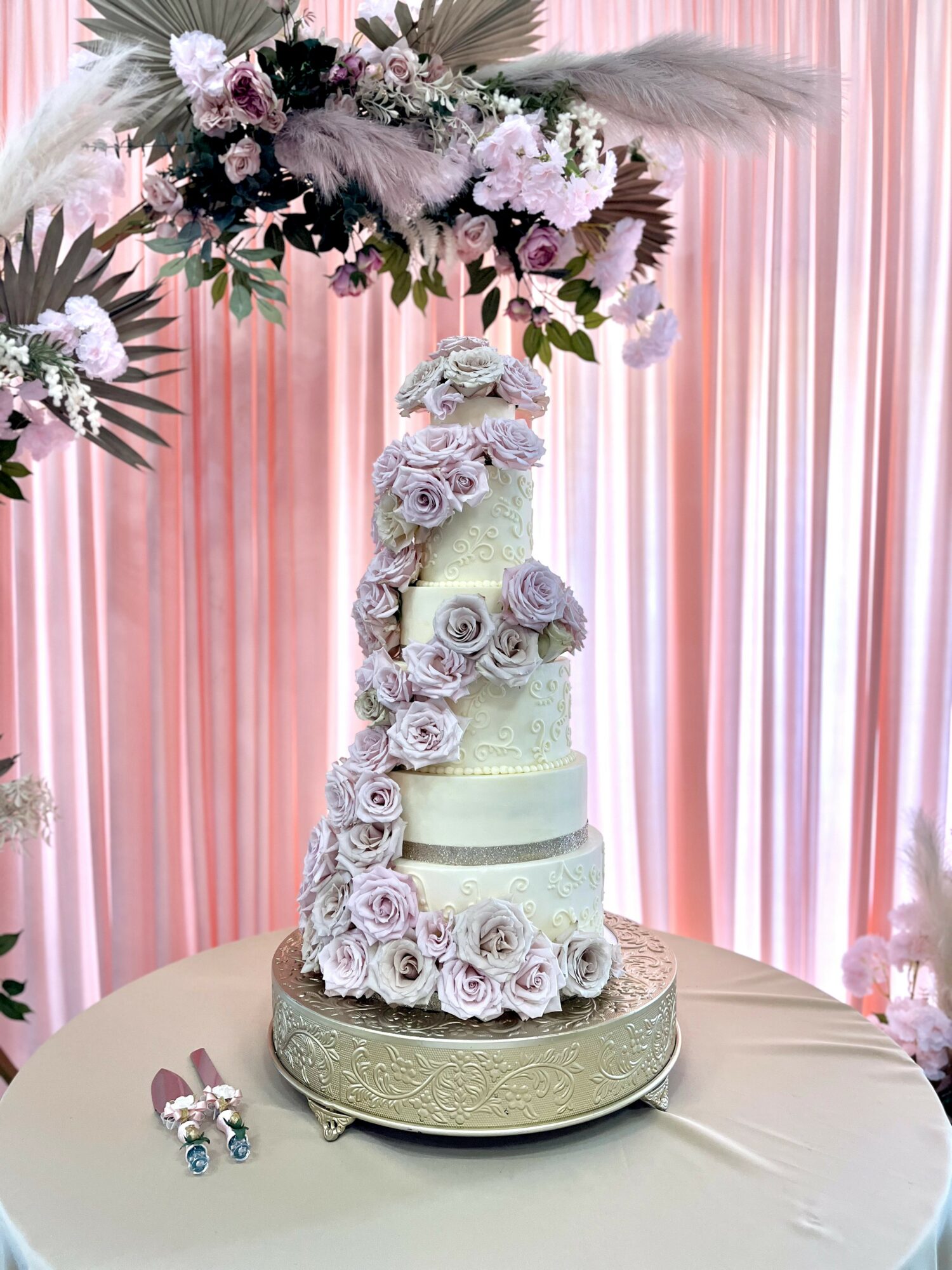 Three-tiered wedding cake decorated with purple roses and white flowers, on a round table with a decorative base, pink curtain backdrop.