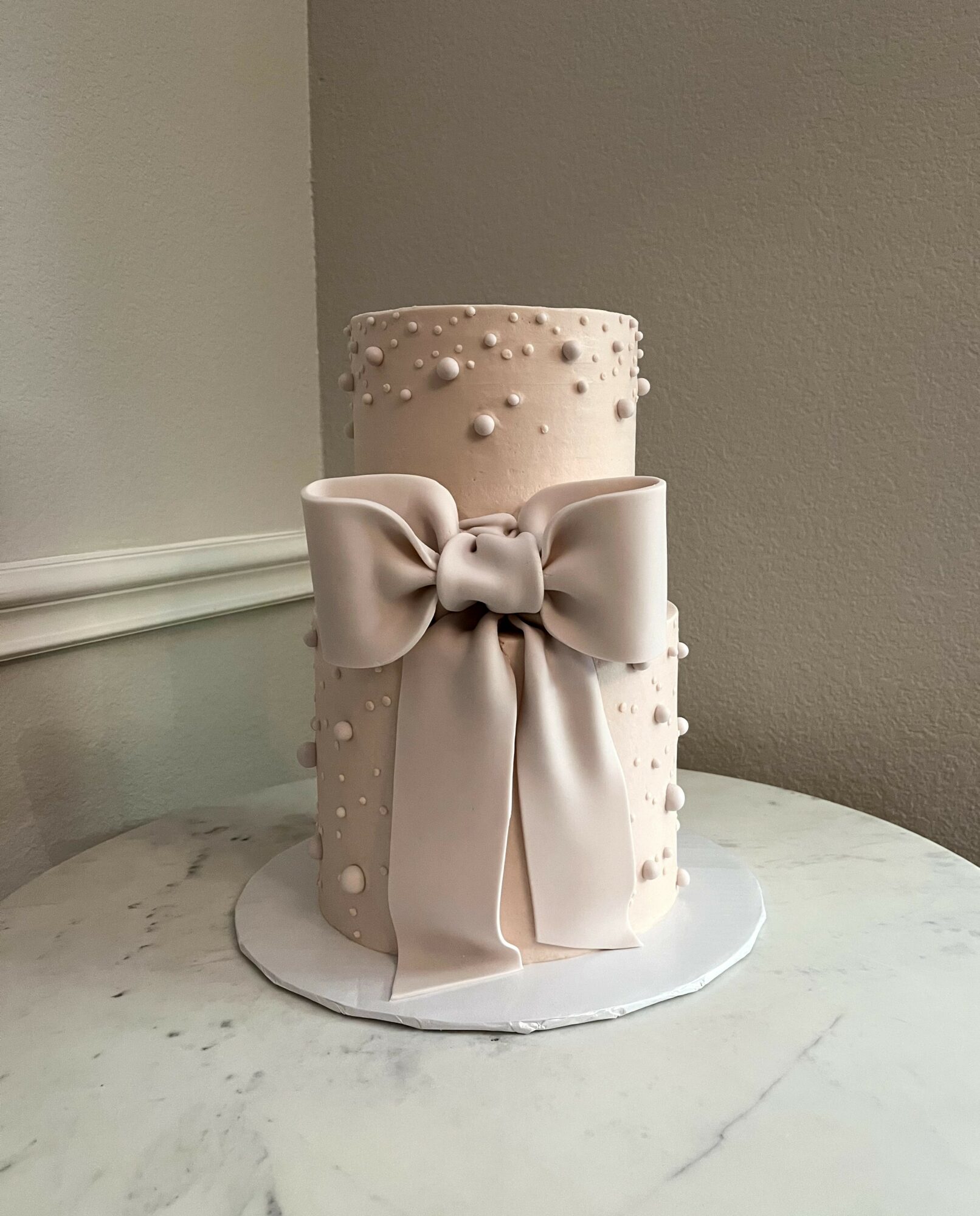 Tall, elegant cake with a large satin bow, decorated with small pearls, on a white marble surface.