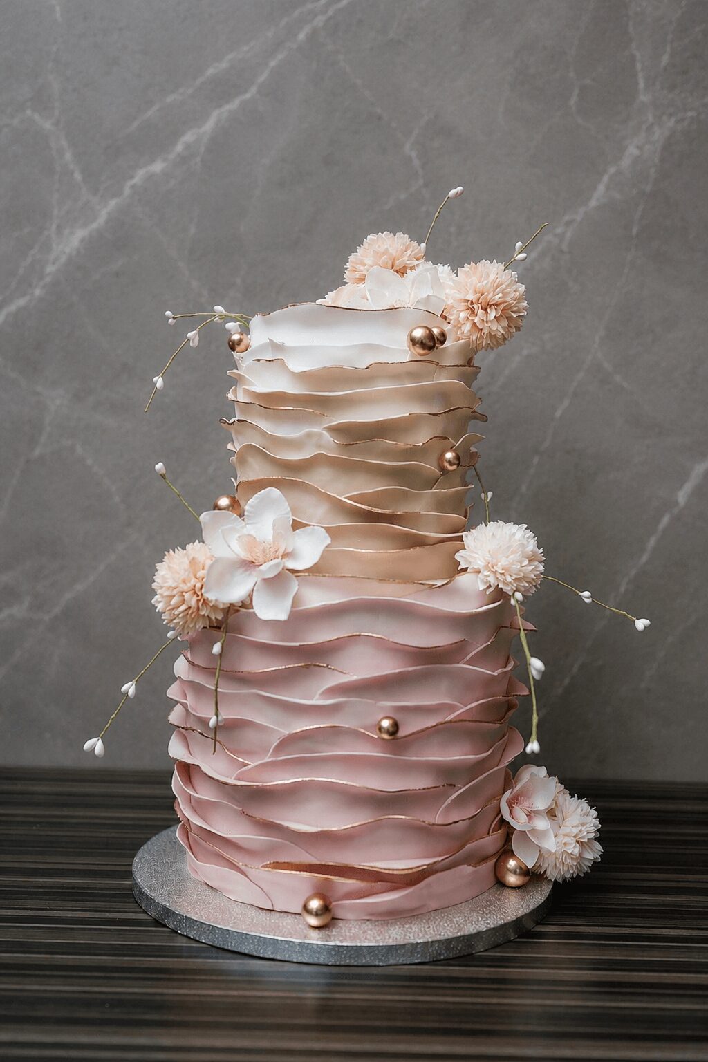 Two-tiered cake with ruffled pink and white icing, decorated with flowers and small gold balls, on a round base.