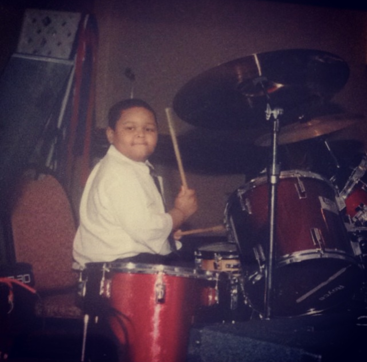 Child playing drums on stage, holding drumsticks, with a drum set and a chair in the background.