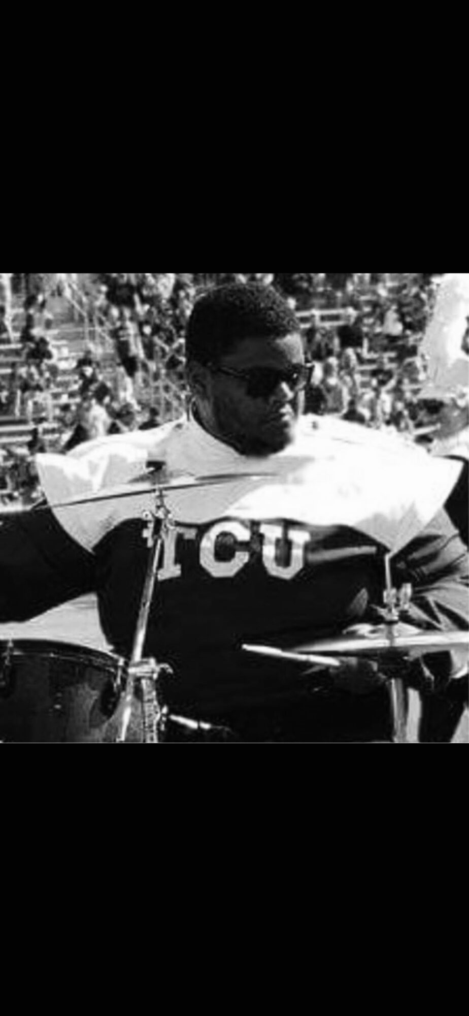 Person wearing sunglasses and a TCU football uniform playing drums outdoors, crowd in background.