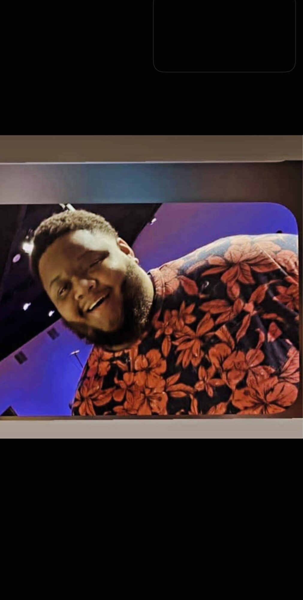 Smiling man with a beard wearing a red floral shirt, indoors with blue lighting, facing the camera.