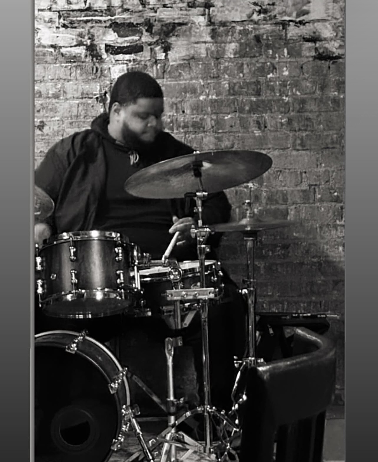 Person playing drums against a brick wall, focused on the drum set, in black and white.