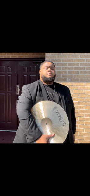 Person holding a cymbal stands outside near a brick wall and dark door, wearing a black jacket and glasses.