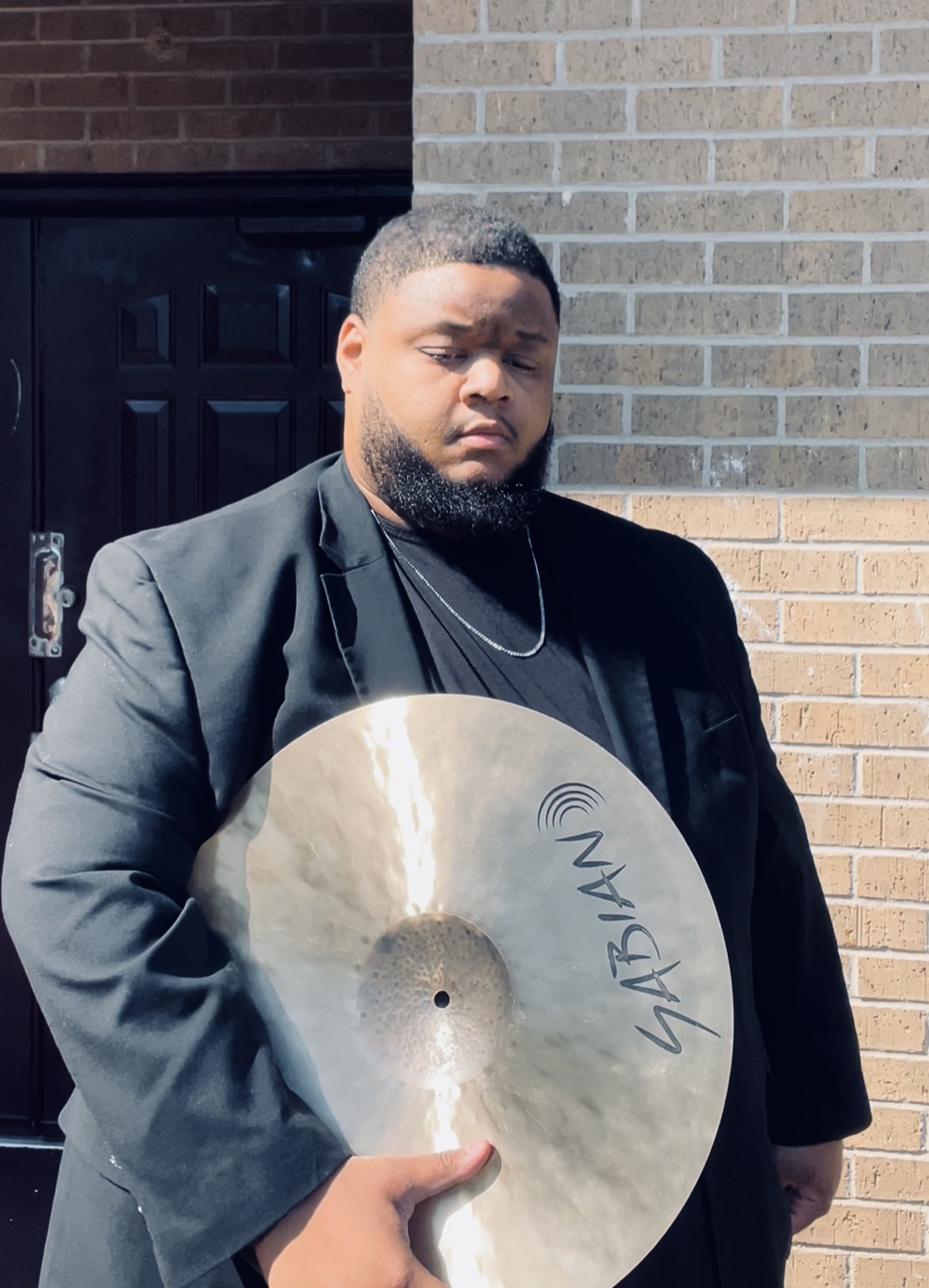 Man with a beard and short hair wearing a black blazer, holding a large cymbal outdoors against a brick wall.