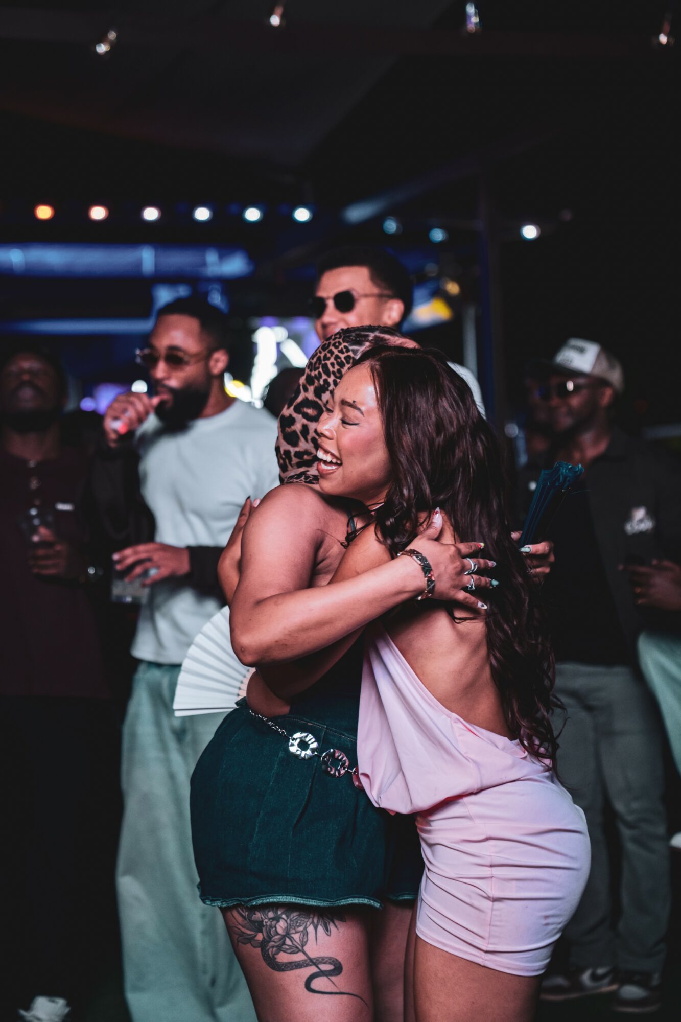 Two women hugging at a party with people and colorful lights in the background.