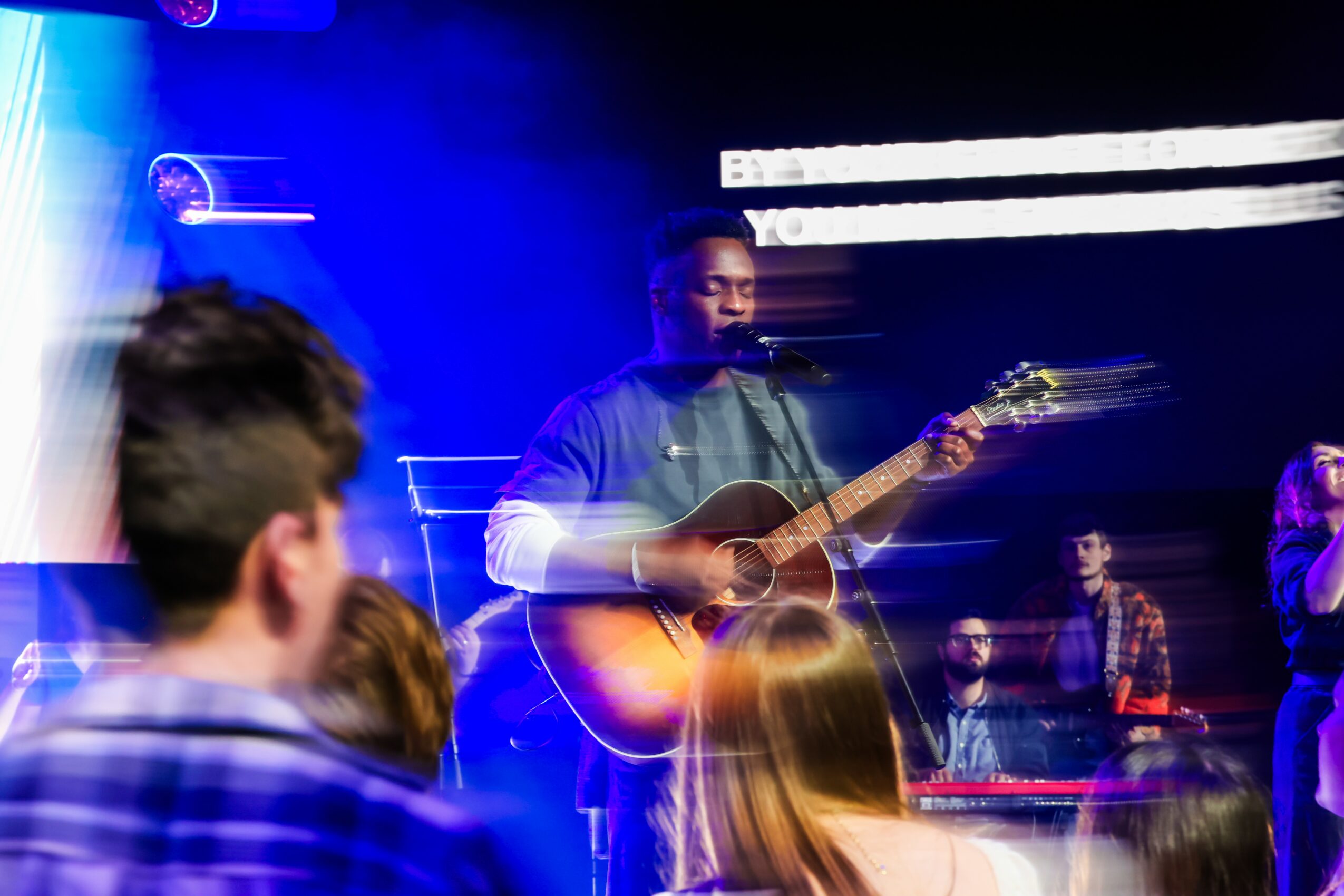 A man plays guitar on stage with a microphone, surrounded by an audience in a dimly lit setting with blue lighting.