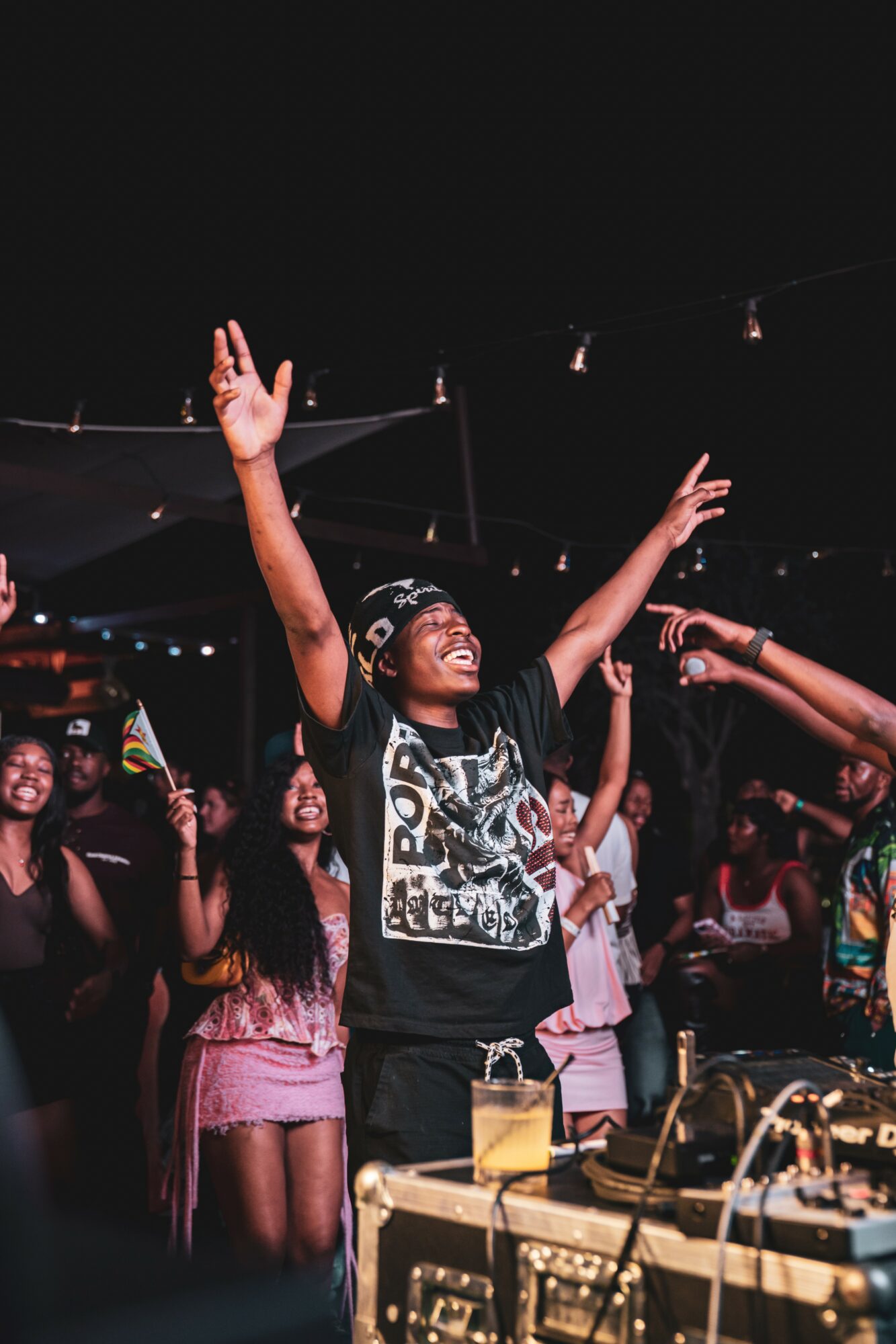 Person with arms raised celebrating at a party with others in the background, DJ equipment visible in foreground.