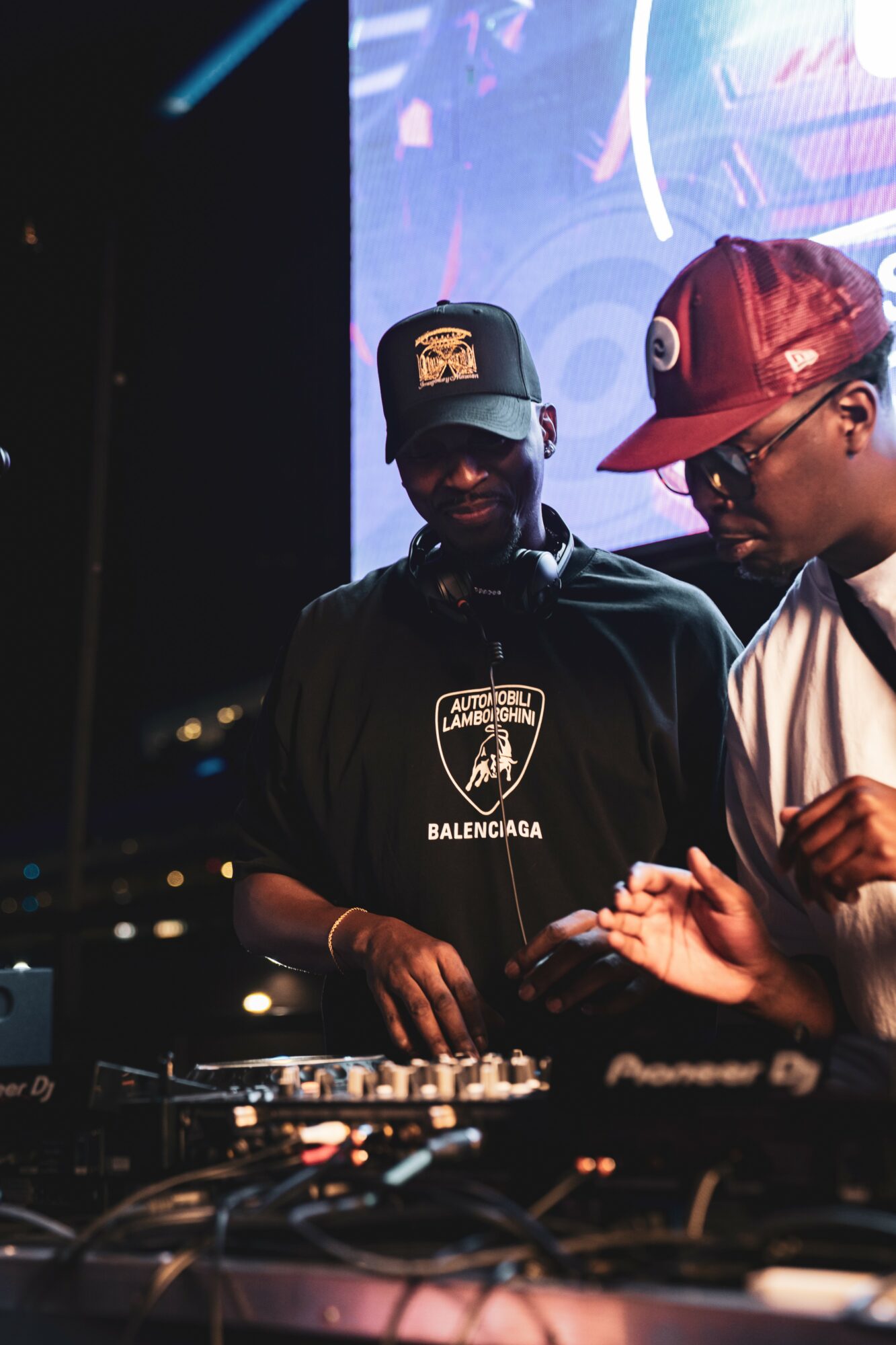 Two DJs perform at a nightclub, one wearing a black cap and shirt, the other in a red cap and glasses, with DJ equipment.