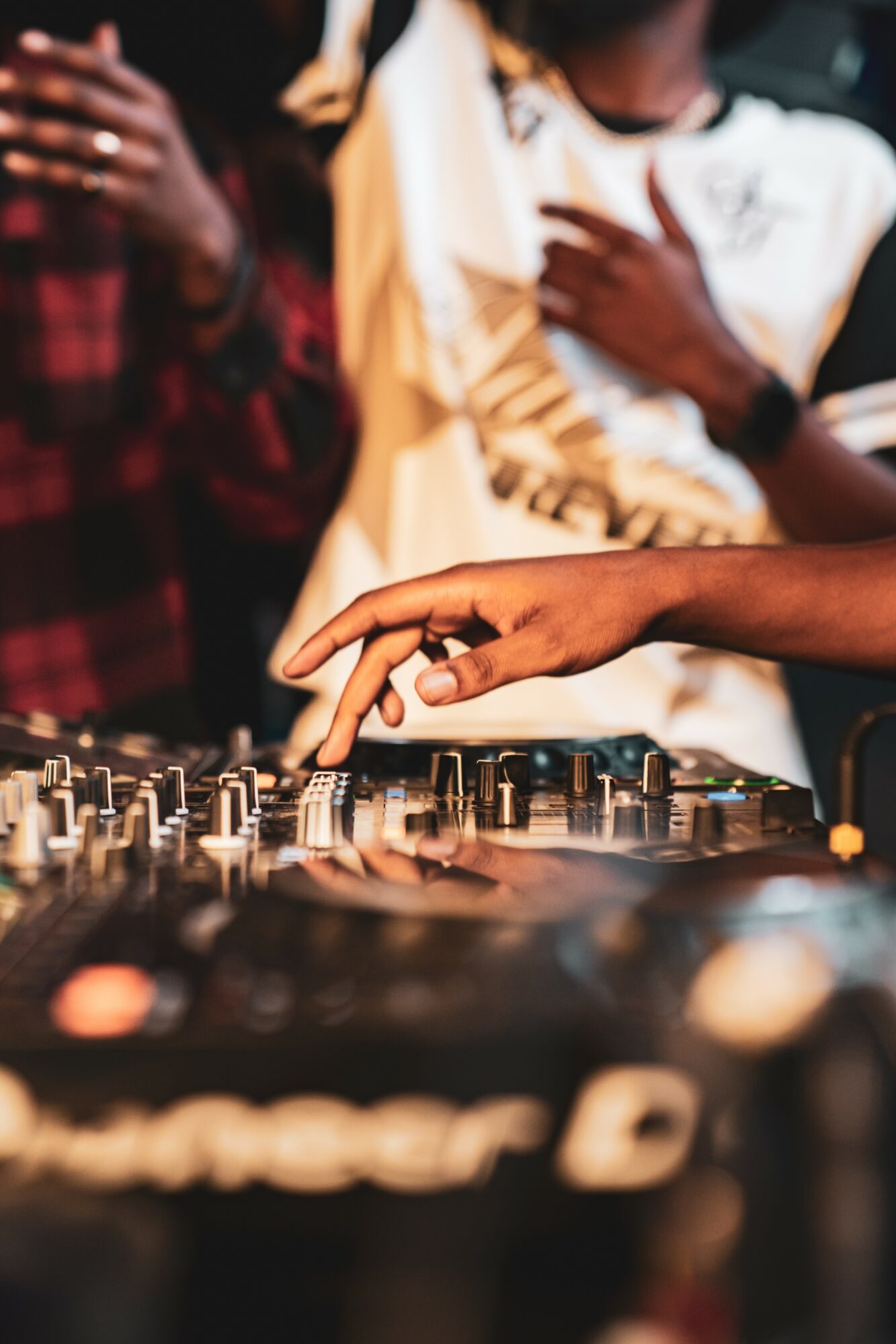 Person's hand adjusting DJ equipment with people in the background, some with crossed arms, in a social setting.