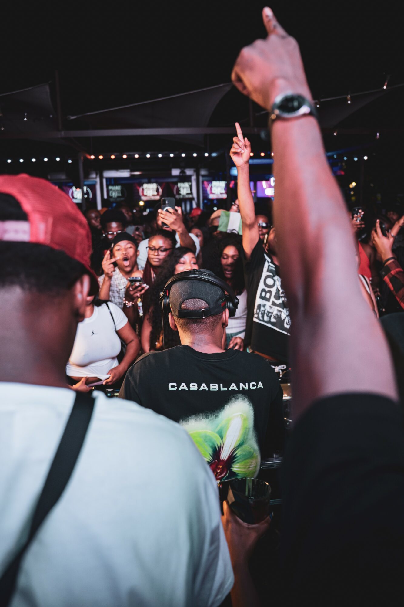 Crowd of people at a concert or event, some raising hands, with a dark background and stage lights.