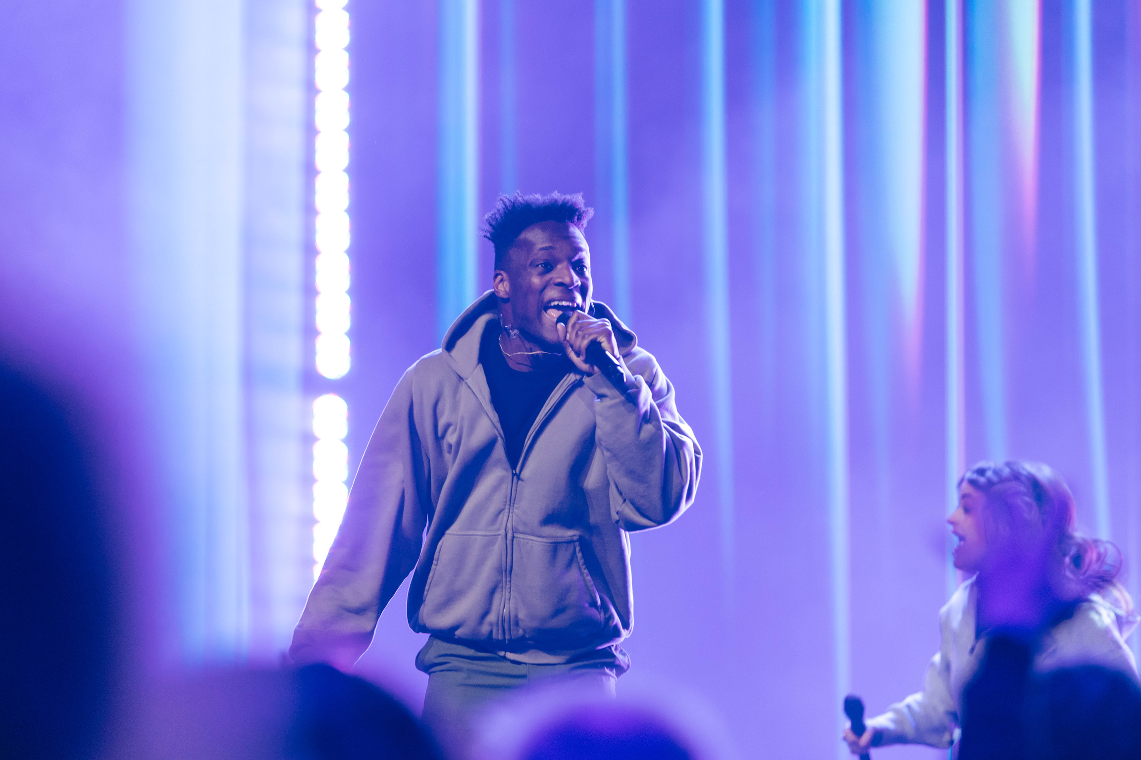 Person singing into a microphone on stage with colorful vertical light beams behind them.