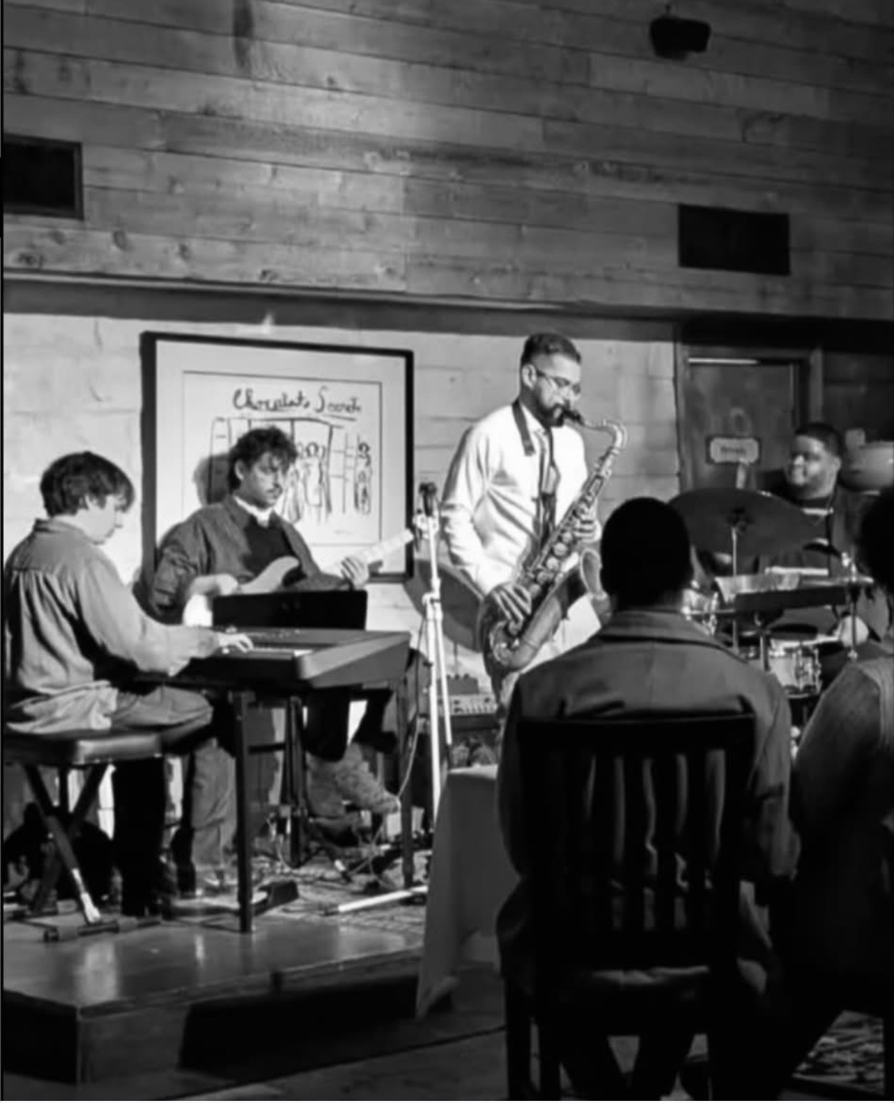 Musicians perform on stage with audience watching, in a room with wooden walls and framed artwork, black and white photo.