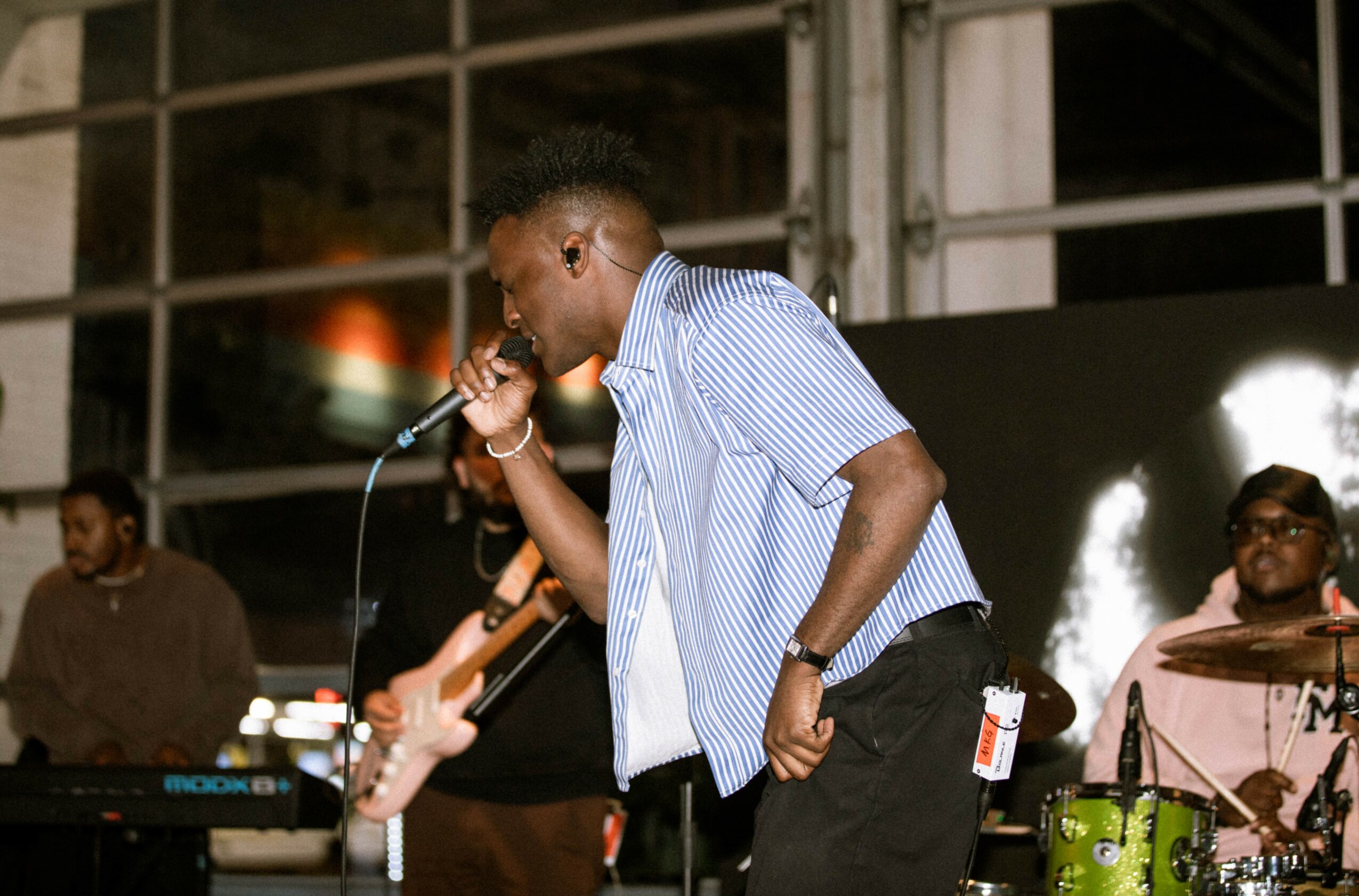 Young man singing into a microphone during a live music performance, with band members playing instruments in the background.
