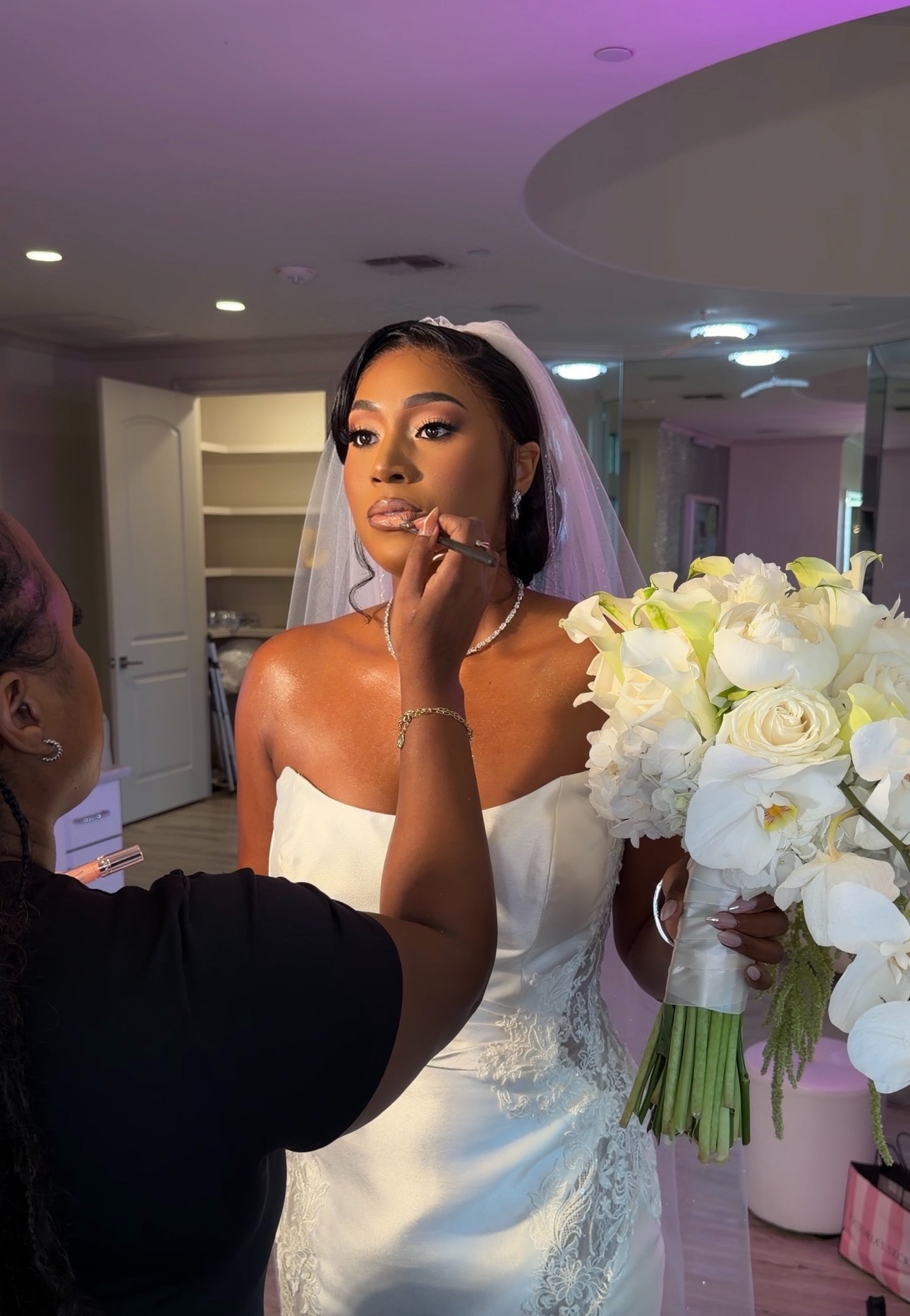 Bride with veil and bouquet being prepared, indoors, with makeup artist applying makeup, soft lighting, elegant setting.