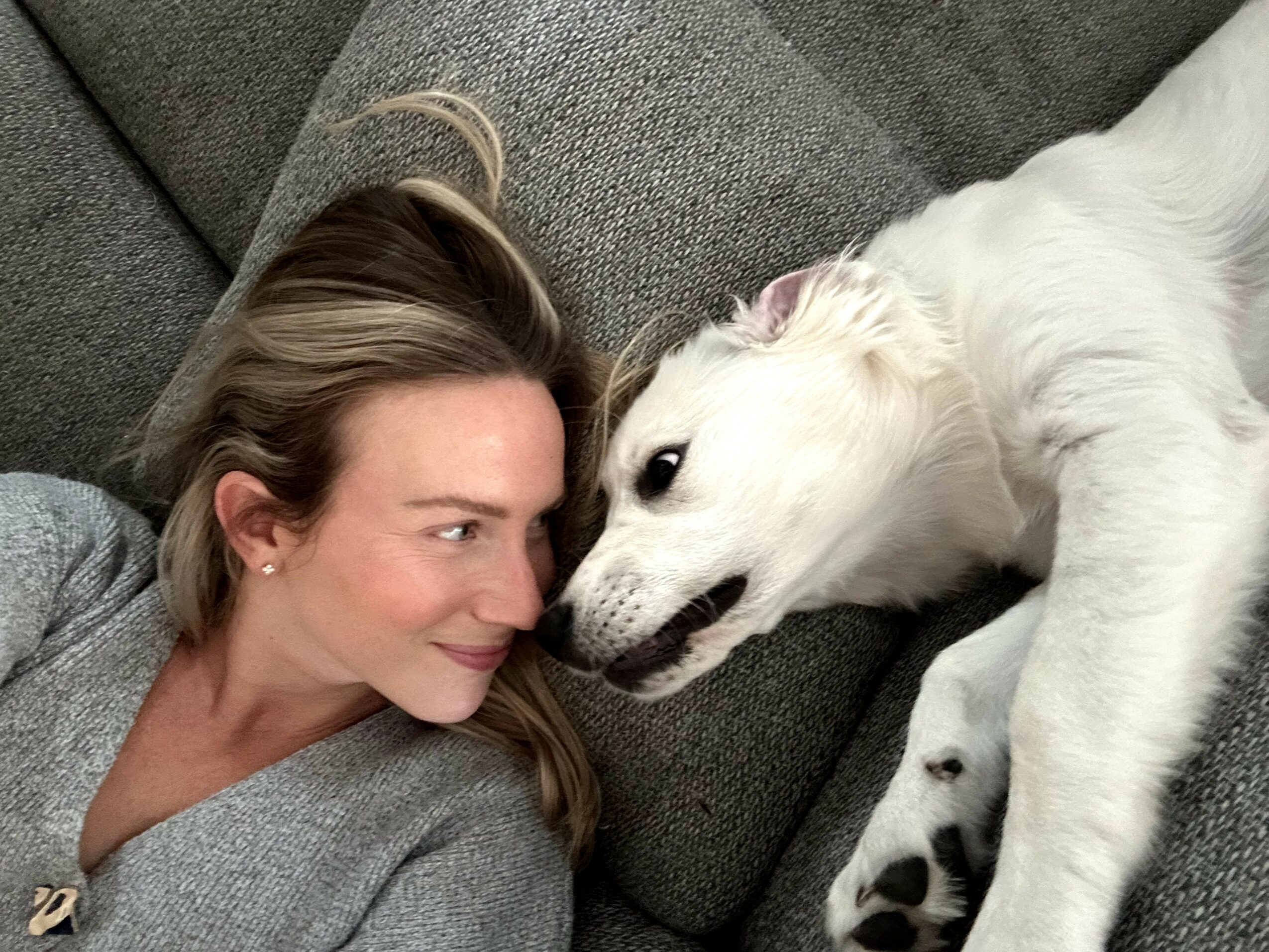 Woman and white dog lying on a gray couch face to face, close together, smiling at each other.