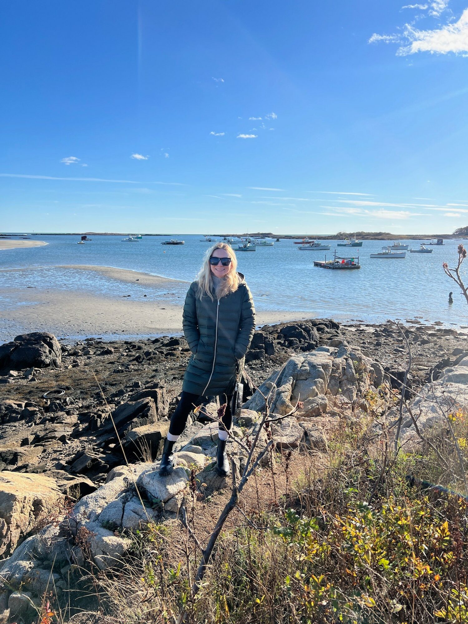 Woman standing on rocky shoreline with boats in water and blue sky in background.