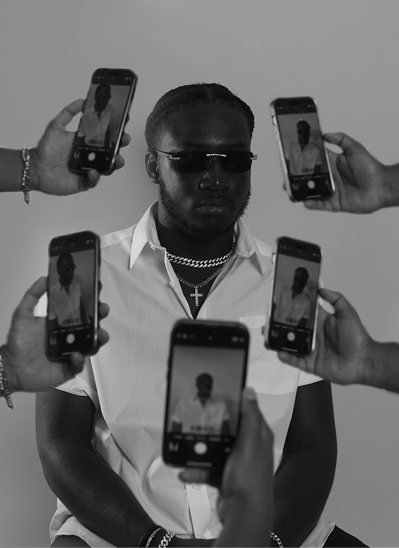Man with sunglasses and jewelry surrounded by people taking photos with smartphones, black and white image.