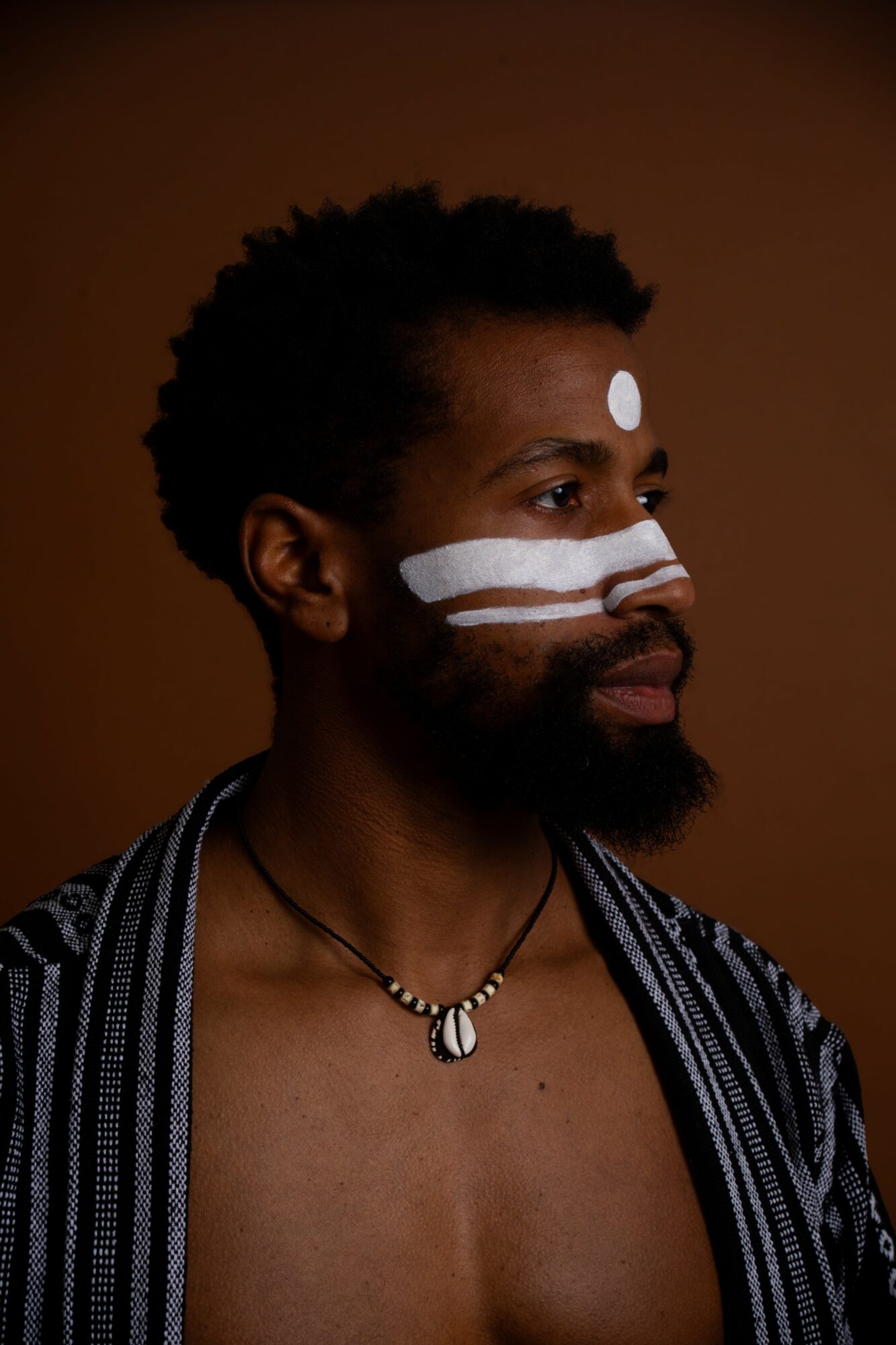 Person with face paint, wearing a striped shirt and necklace, against a brown background.