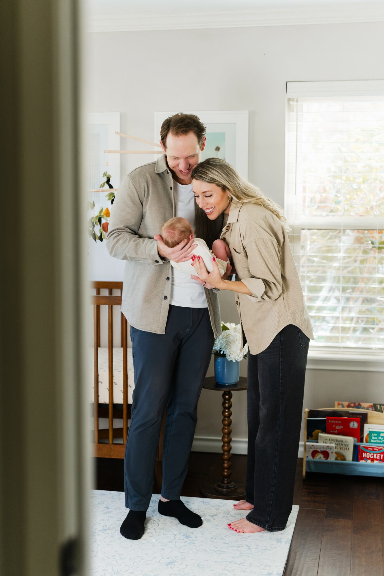 A couple holding a newborn baby in a bright room with large windows, a wooden crib, and books on a shelf.