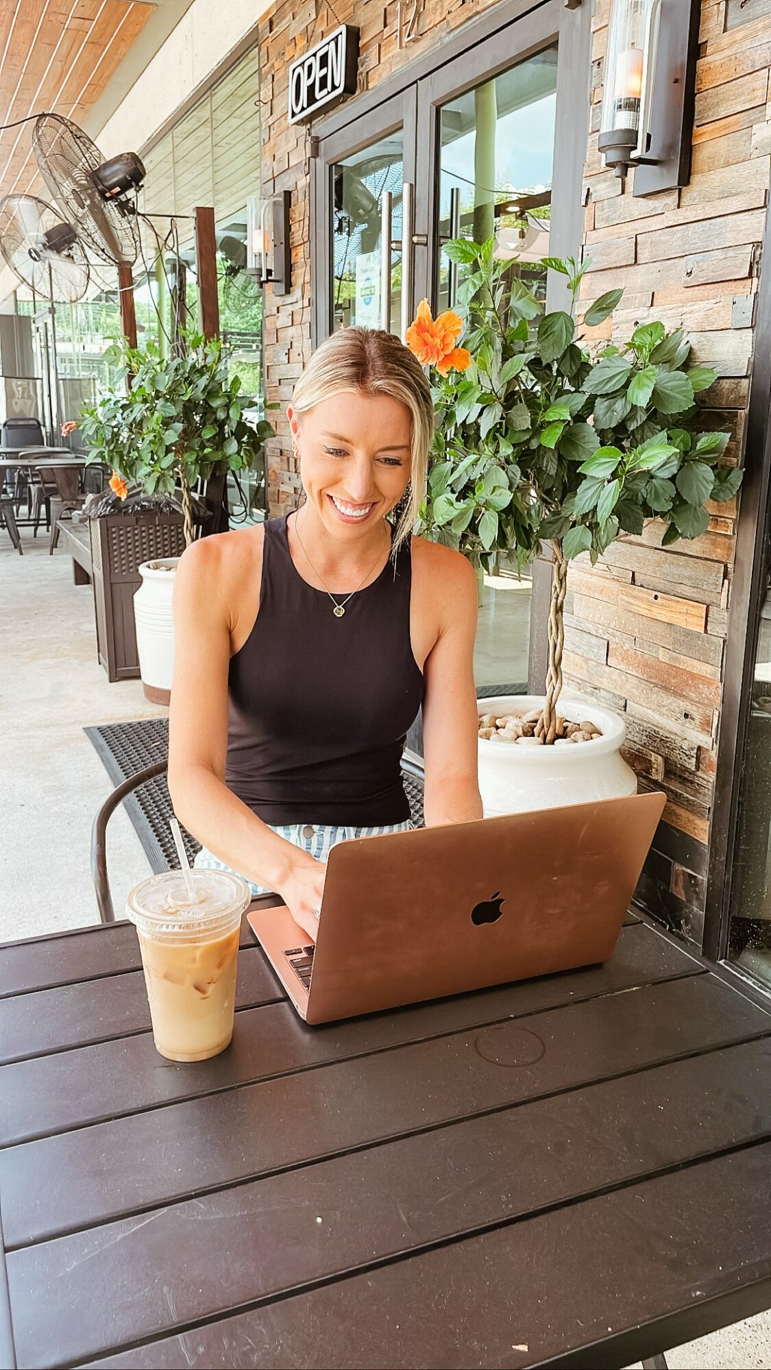 Woman sitting at outdoor table with laptop and iced coffee, smiling, plants and brick wall in background.