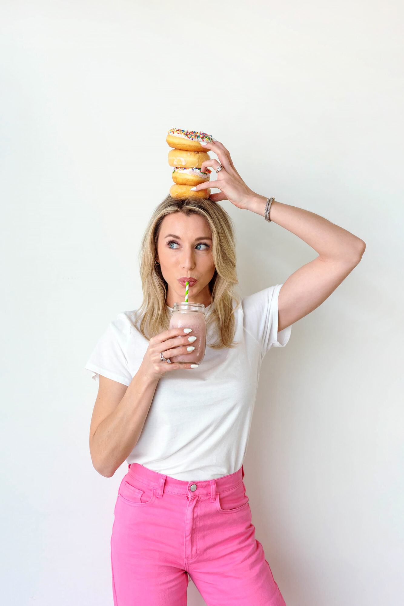 Woman with blonde hair in a white shirt and pink pants holds a drink and balances a stack of donuts on her head.