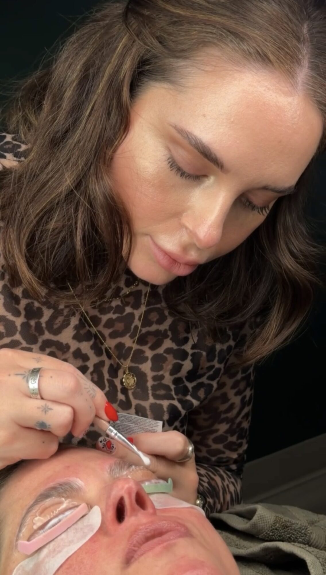 Woman with long brown hair looking down at a person receiving a cosmetic treatment on their face, wearing a leopard print top.