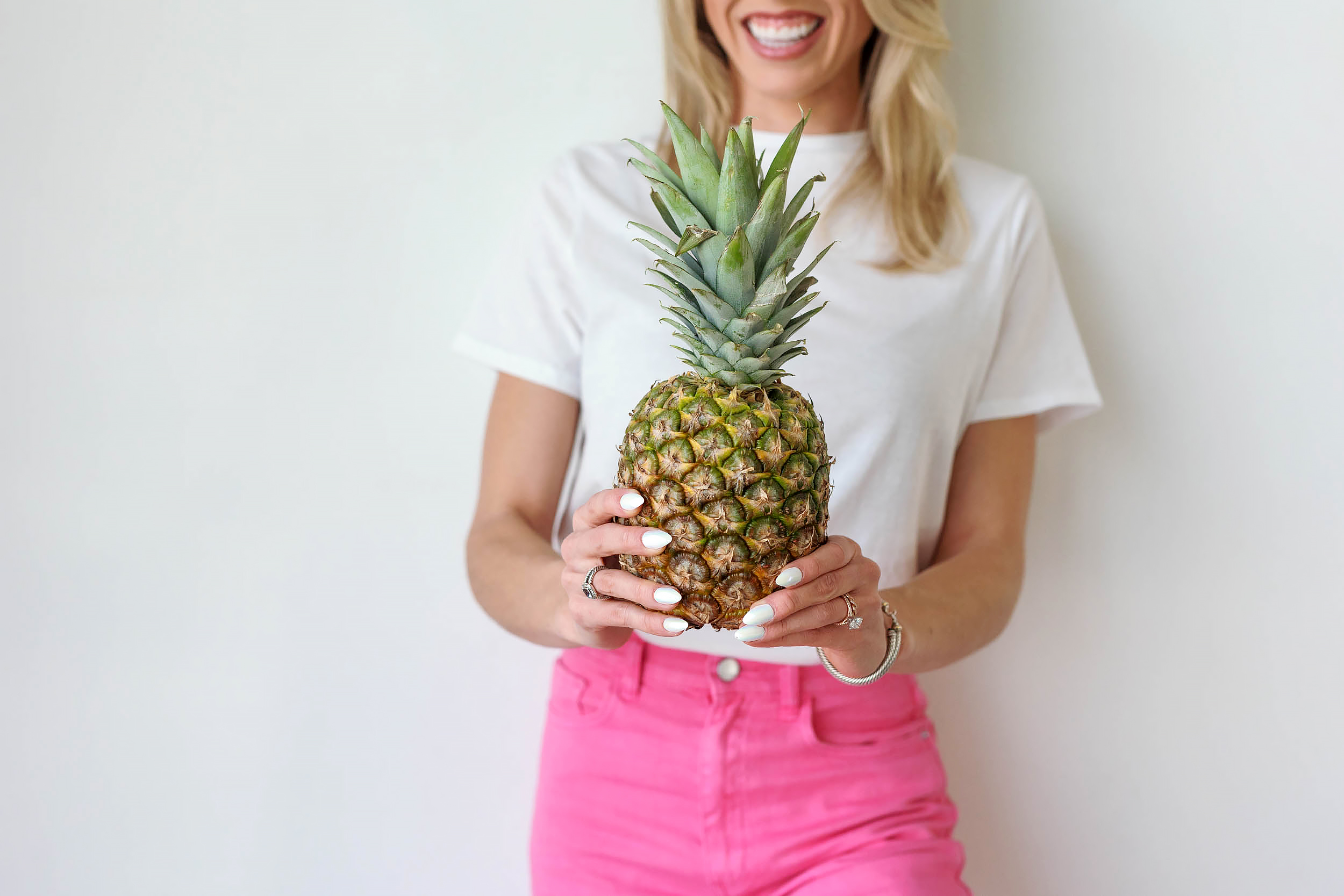Person holding a pineapple, smiling, wearing a white shirt and pink pants, against a plain background.
