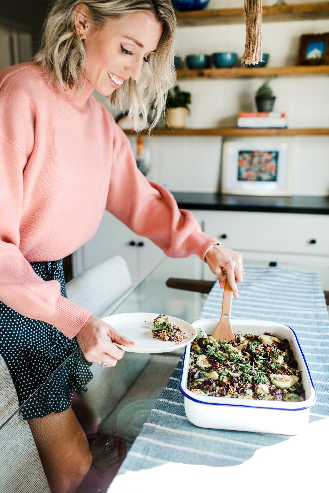 Woman smiling and serving food from a baking dish in a bright kitchen, wearing a pink sweater and dark skirt.