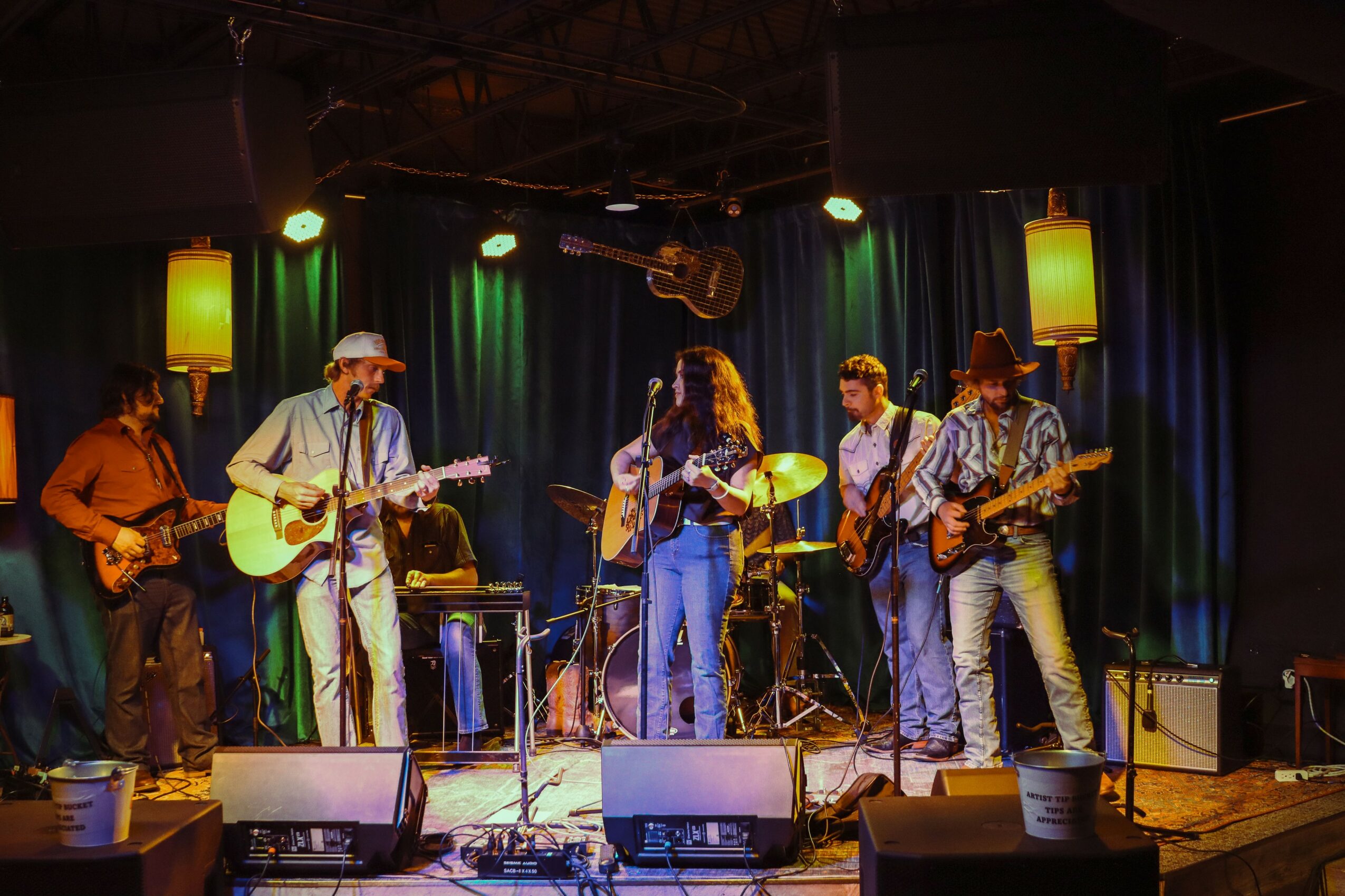 Five musicians perform on stage with guitars and drums, colorful stage lighting, and a dark curtain backdrop.