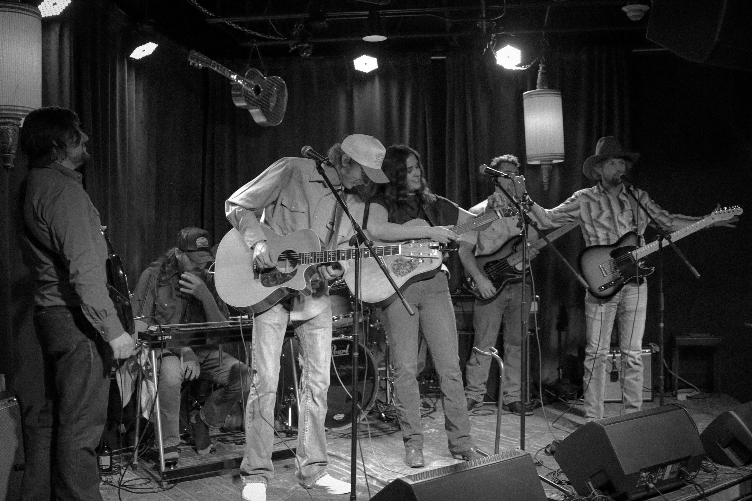 Group of six musicians performing on stage with guitars and drums, stage lighting, and a dark curtain backdrop.