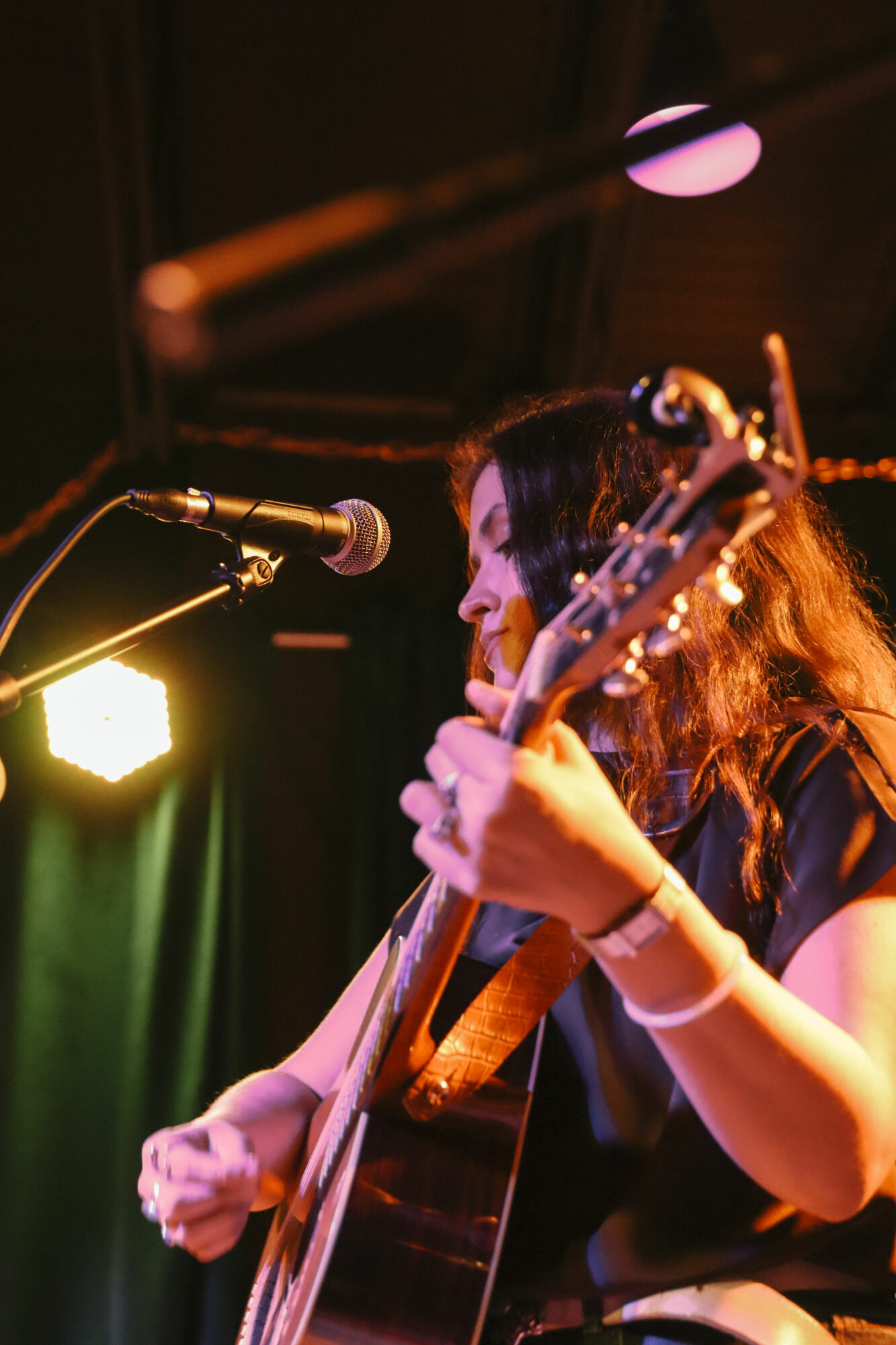 Woman playing guitar and singing into microphone on stage with colorful lights.