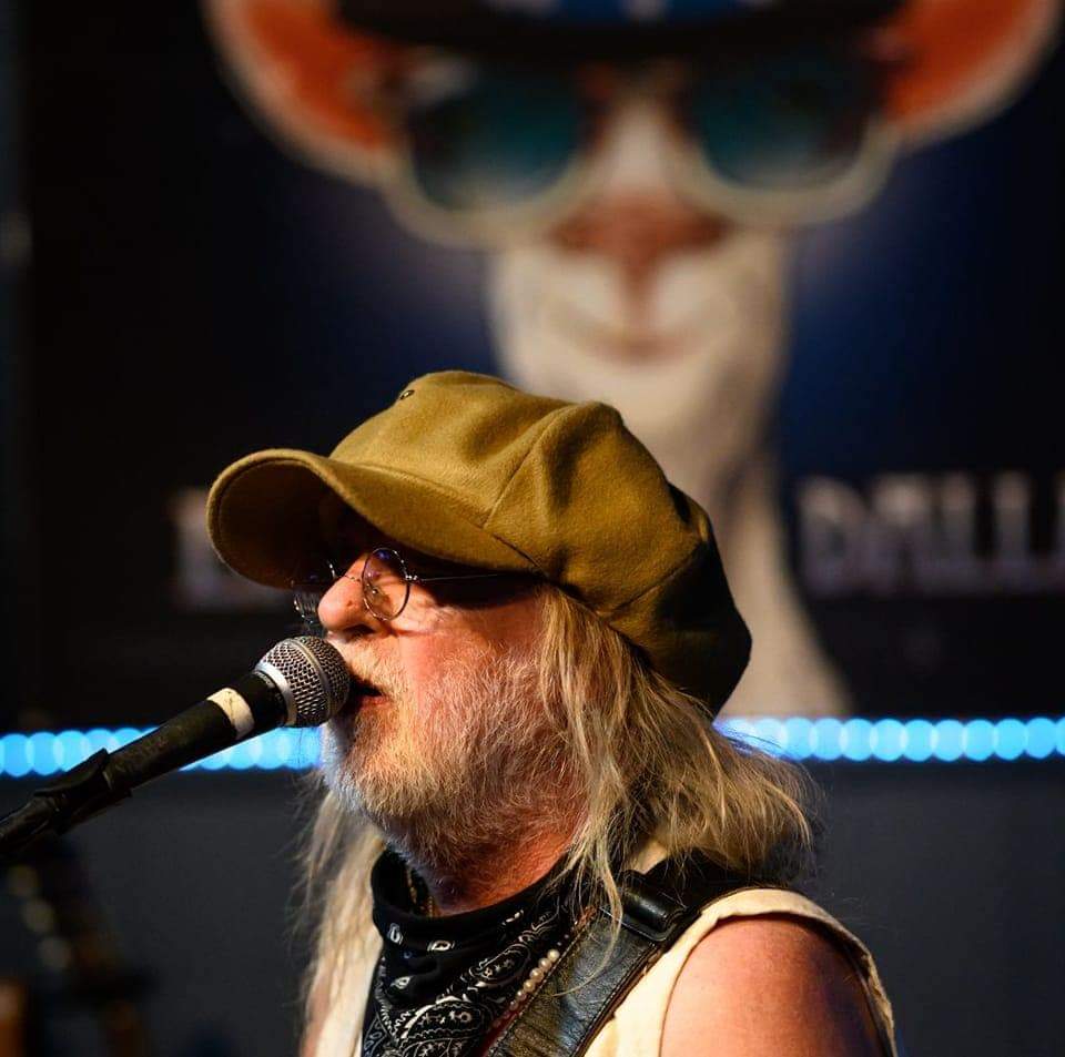 A man with long hair, beard, glasses, and a hat singing into a microphone, with a blurred face in the background.