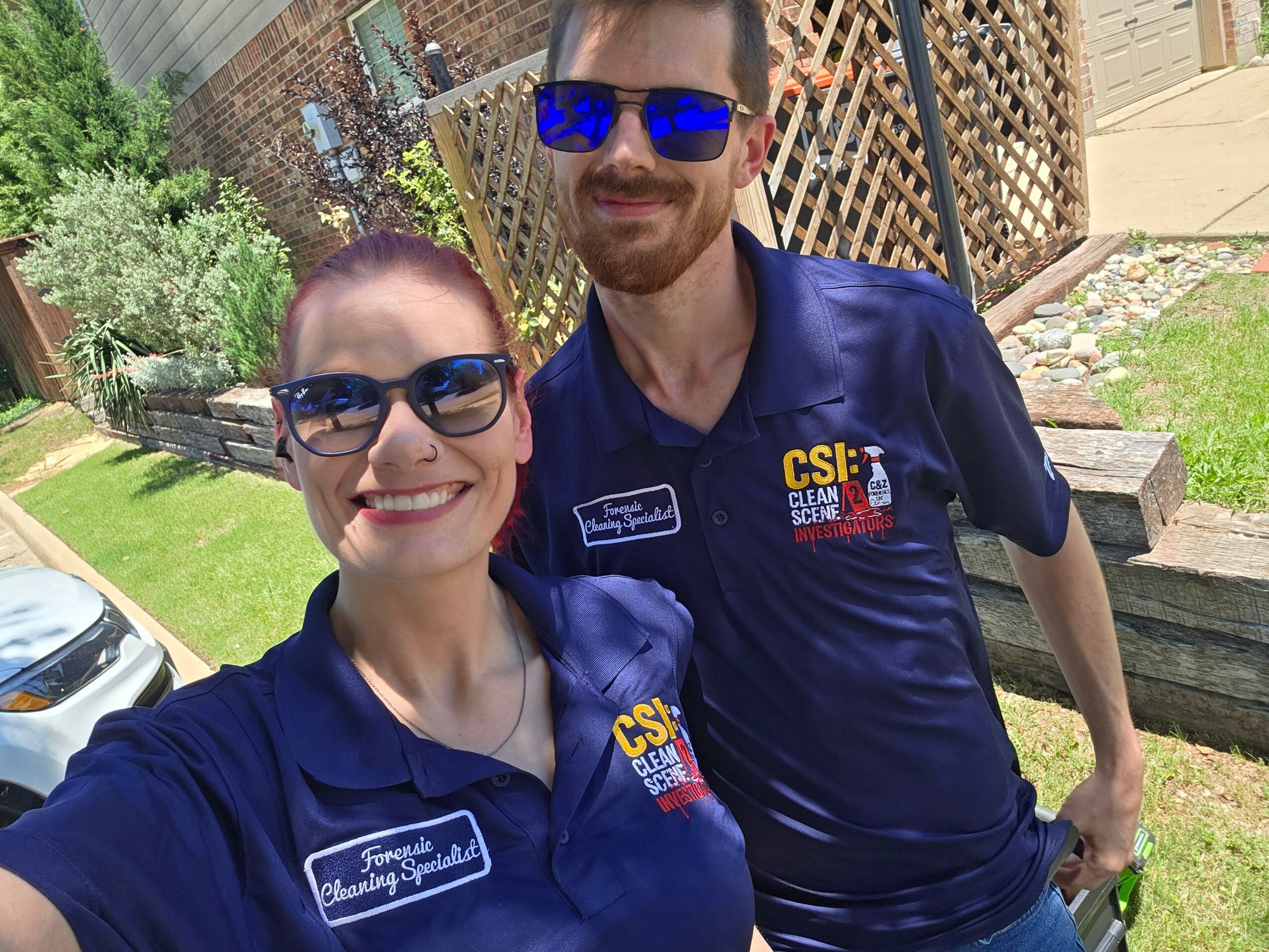 Two people wearing blue shirts and sunglasses outdoors, smiling, with a garden and wooden fence in the background.