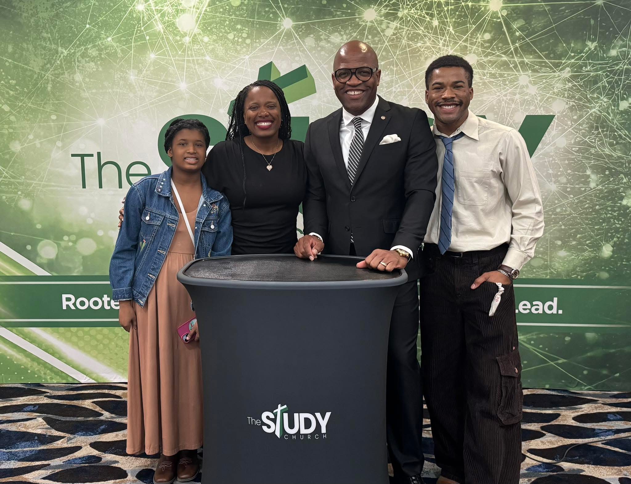 Four people standing behind a table with a green background and text, smiling for a photo.