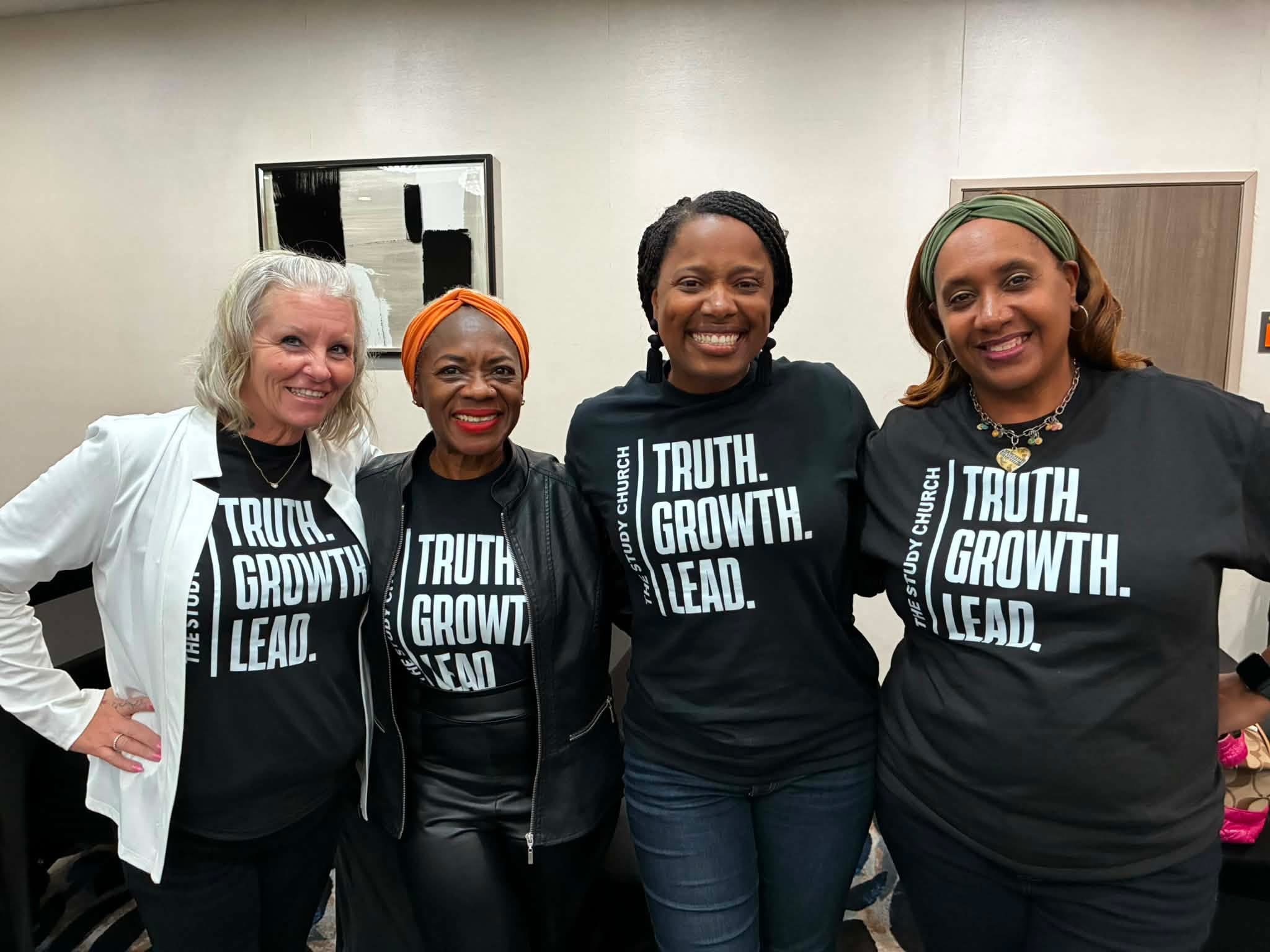 Four women standing together indoors, smiling, wearing black shirts with white text, and one woman in a white jacket, in a room with mirrors and a door.