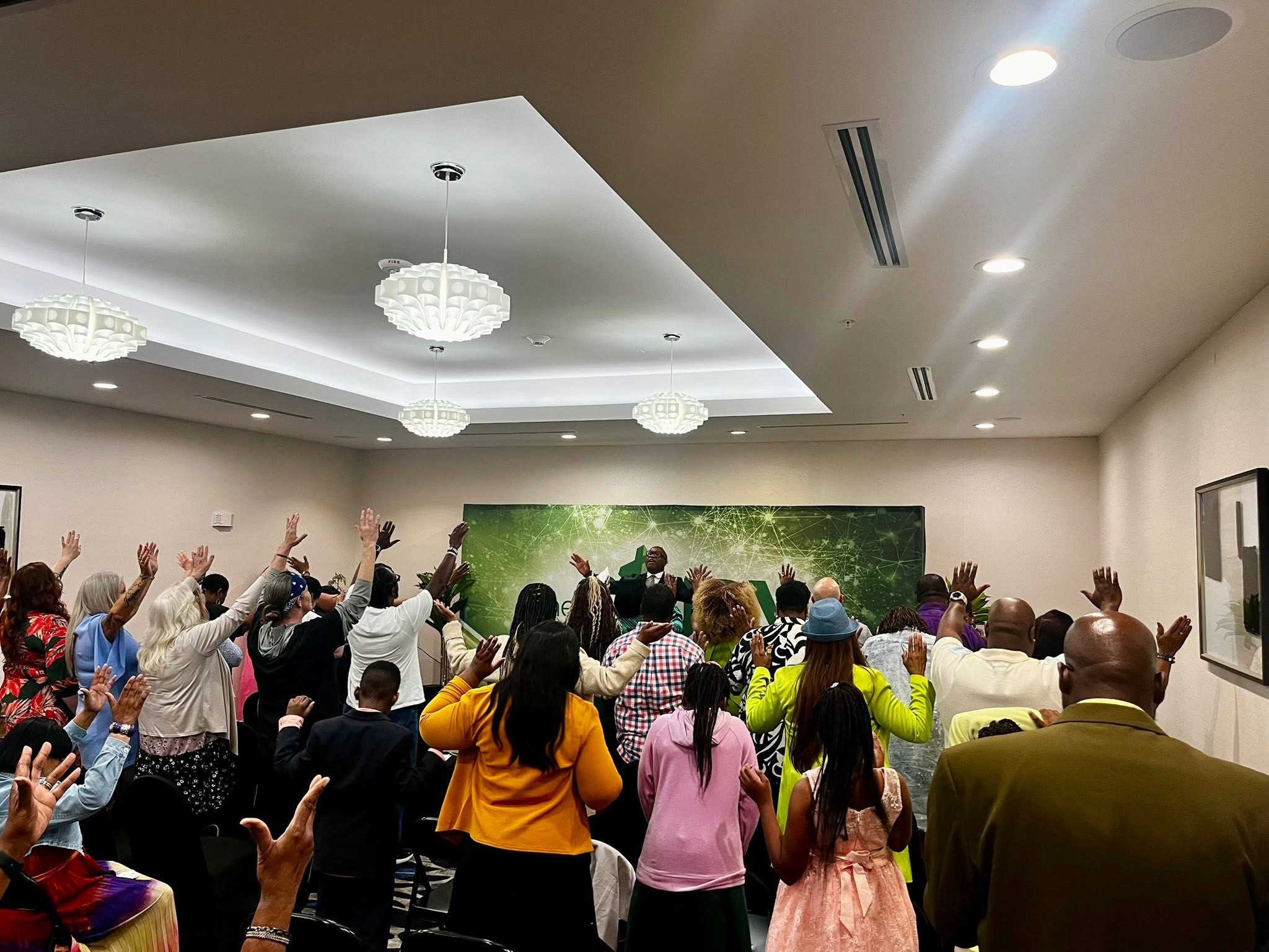 Group of people raising hands in a room with a green backdrop, ceiling lights, and framed pictures on the wall.