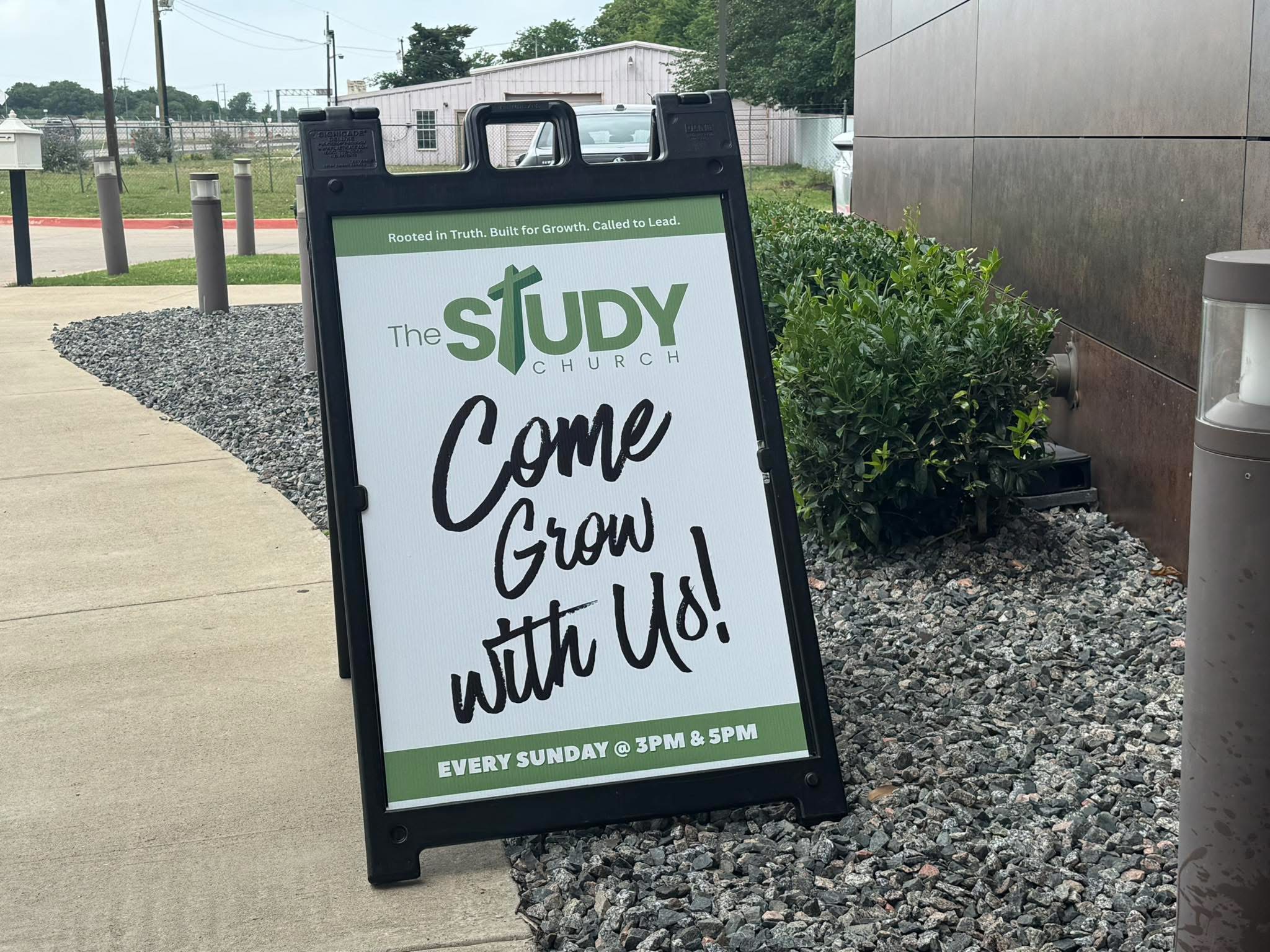 A black A-frame signboard on a sidewalk advertising a church event, with text inviting people to come grow with us, every Sunday at 3 PM and 5 PM.